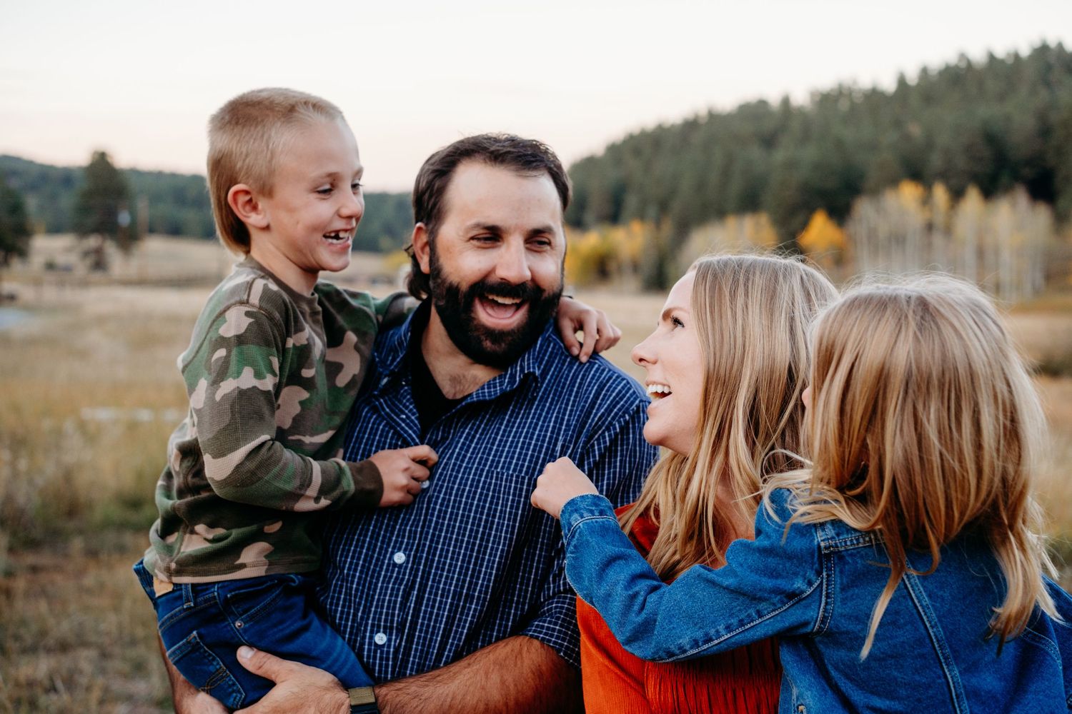 Family of four standing closely together, all smiling and laughing at one another, fall foliage in background.