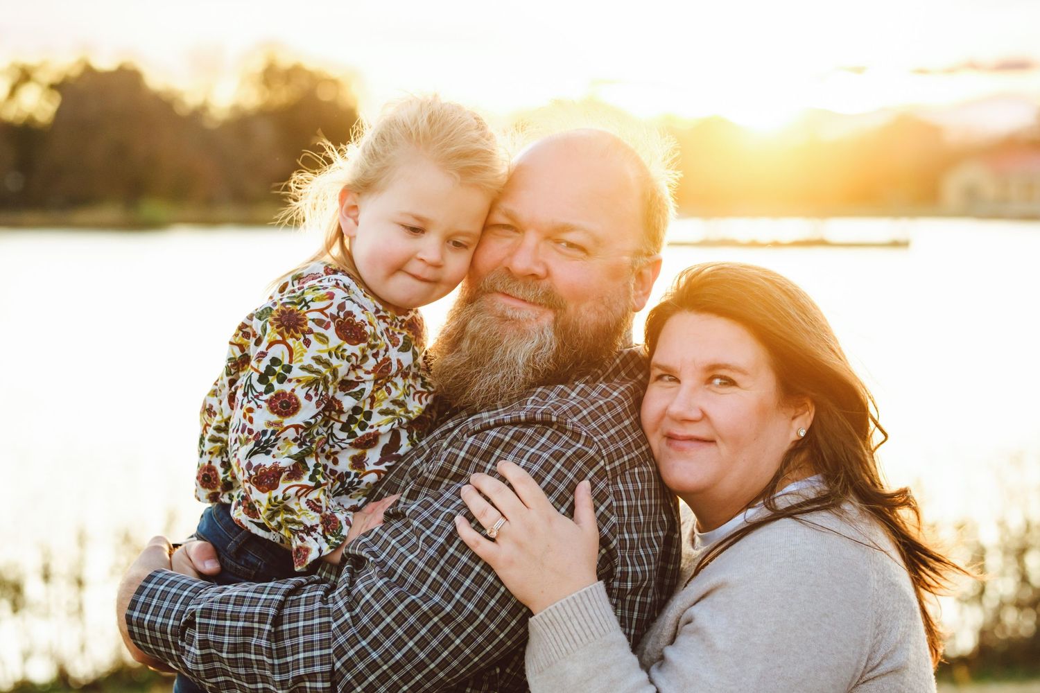 Close up of father holding toddler daughter and mother standing behind husband, holding on to his shoulder, lake in background.