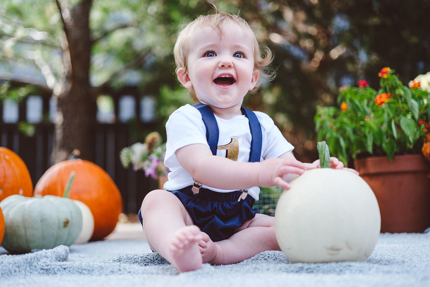 One year old boy sitting next to white pumpkin while sitting on a blanket in his backyard.