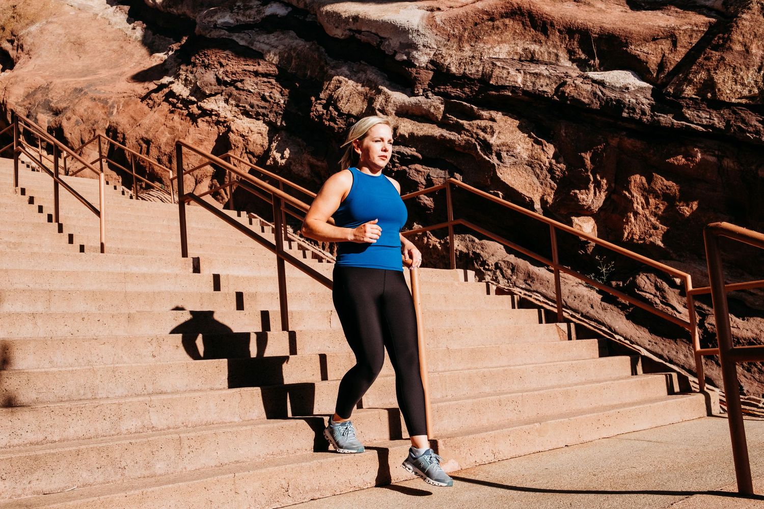 Female trainer running down steps at Red Rocks Amphitheater on a sunny day with harsh shadows.