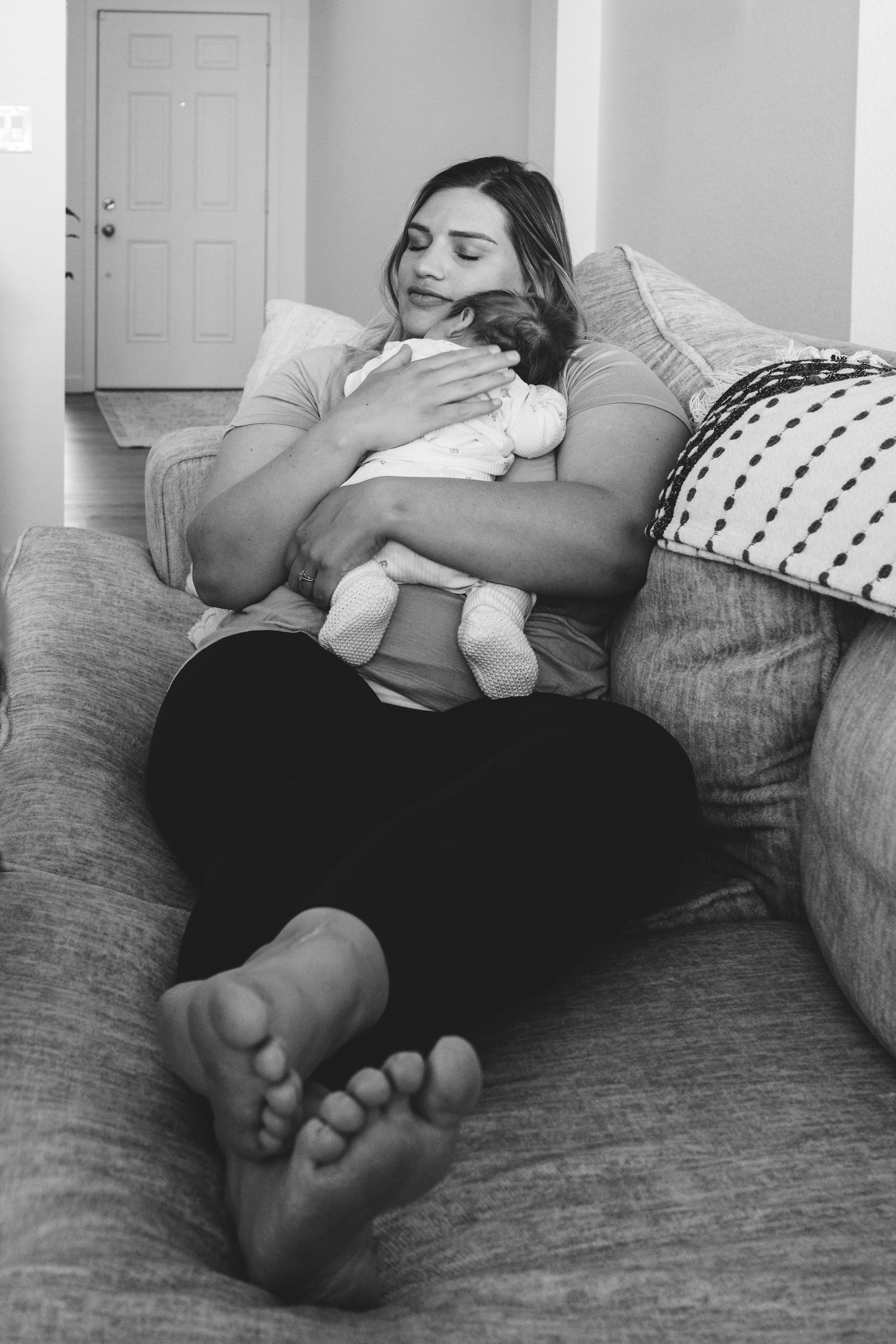 Black and white portrait of mother snuggling newborn son on her chest, legs outstretched in front of her.