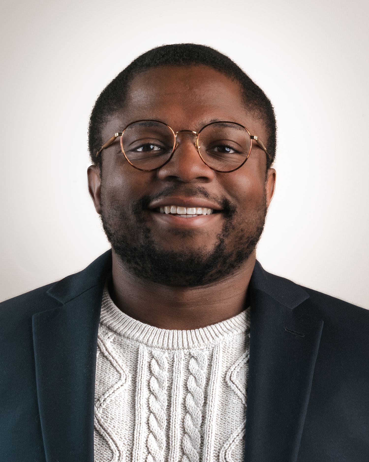 Professional headshot of adult, Black male, wearing glasses, knit sweater and black blazer.