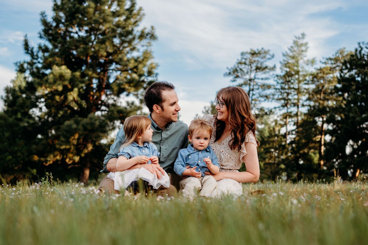 Mother and father sitting in field, each with a child in their lap, surrounded by green grass, tall evergreens and blue sky with fluffy clou