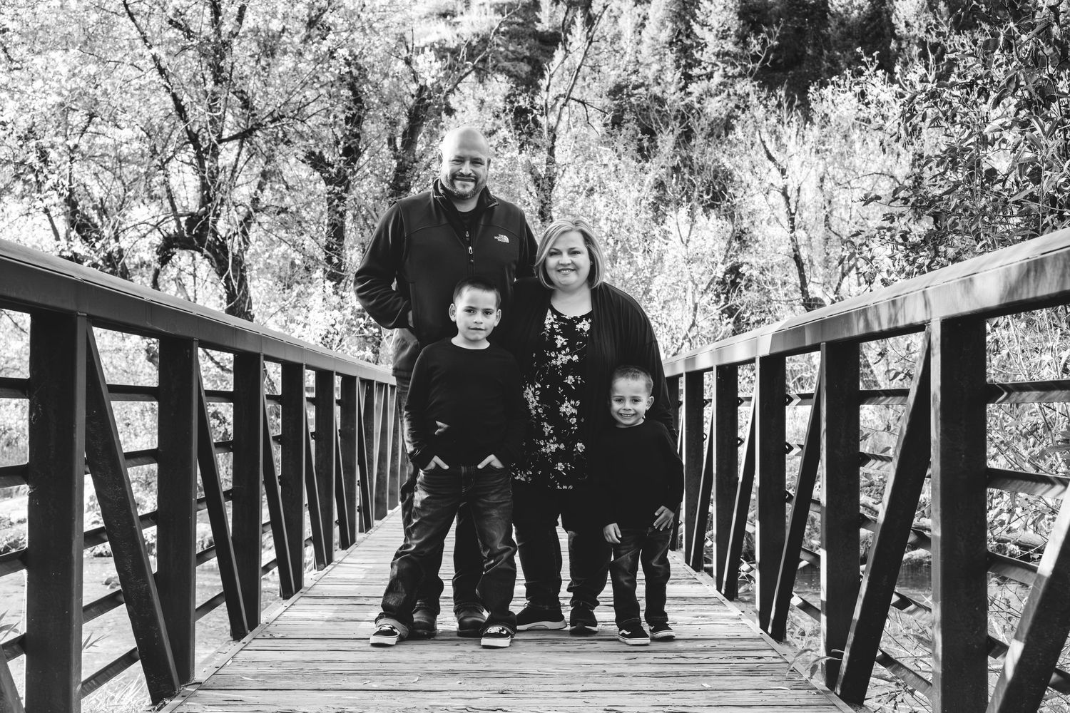 Family of four standing in middle of wooden bridge, iron railing on sides.