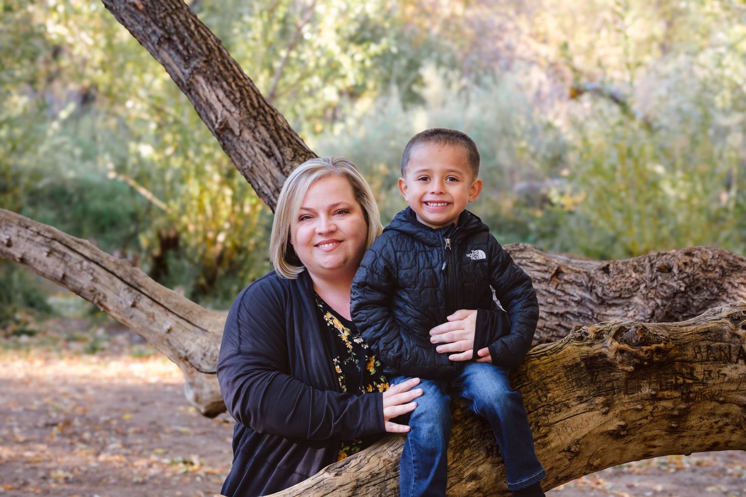 Young boy sitting on tree limb, mother standing behind him, both smiling. Greenery in background.