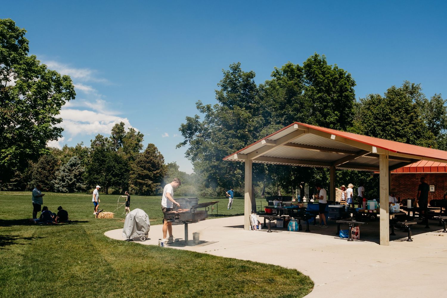 Man grilling at park near shelter with picnic benches, family sitting under park shelter and under shade of trees.