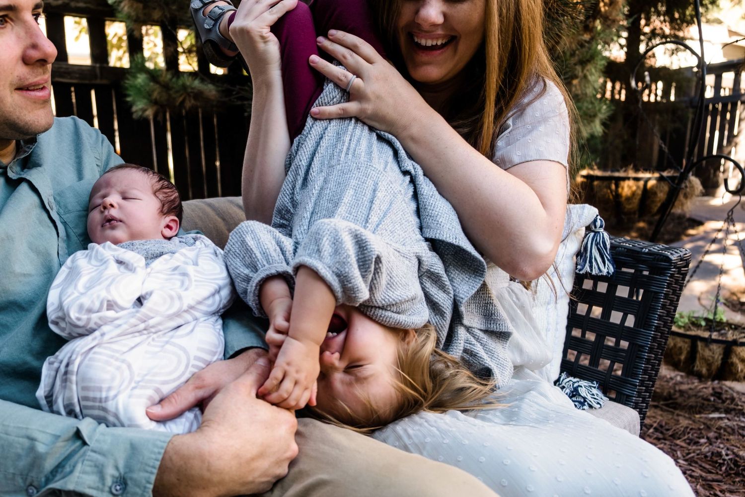 Father holding newborn son, mother holding toddler daughter upside down, tickling her, toddler daughter laughing.