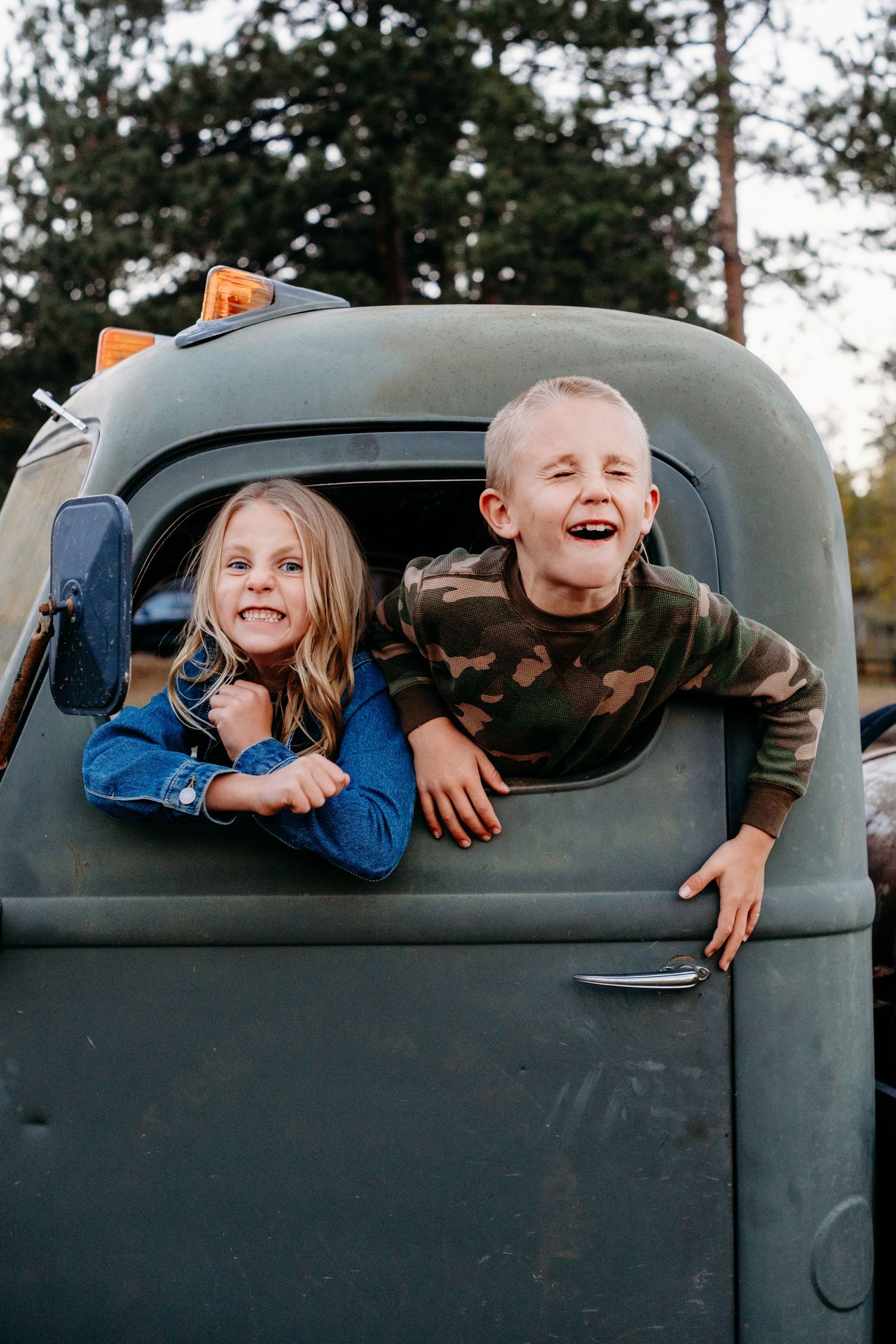 Brother and sister playfully sticking their heads out the window of a vintage green truck.