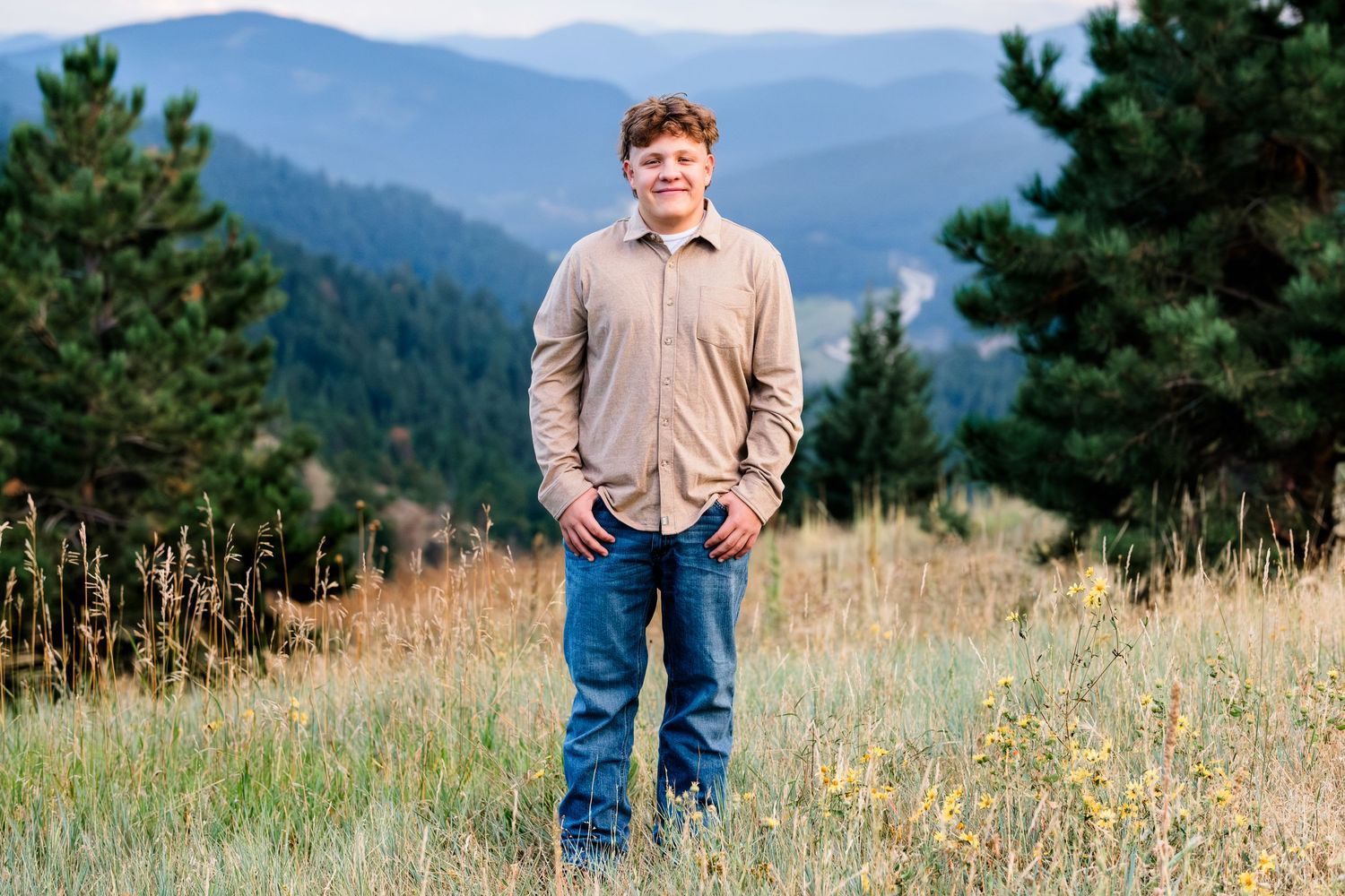 Senior portrait of teen boy standing in field, evergreen trees and mountains in background.