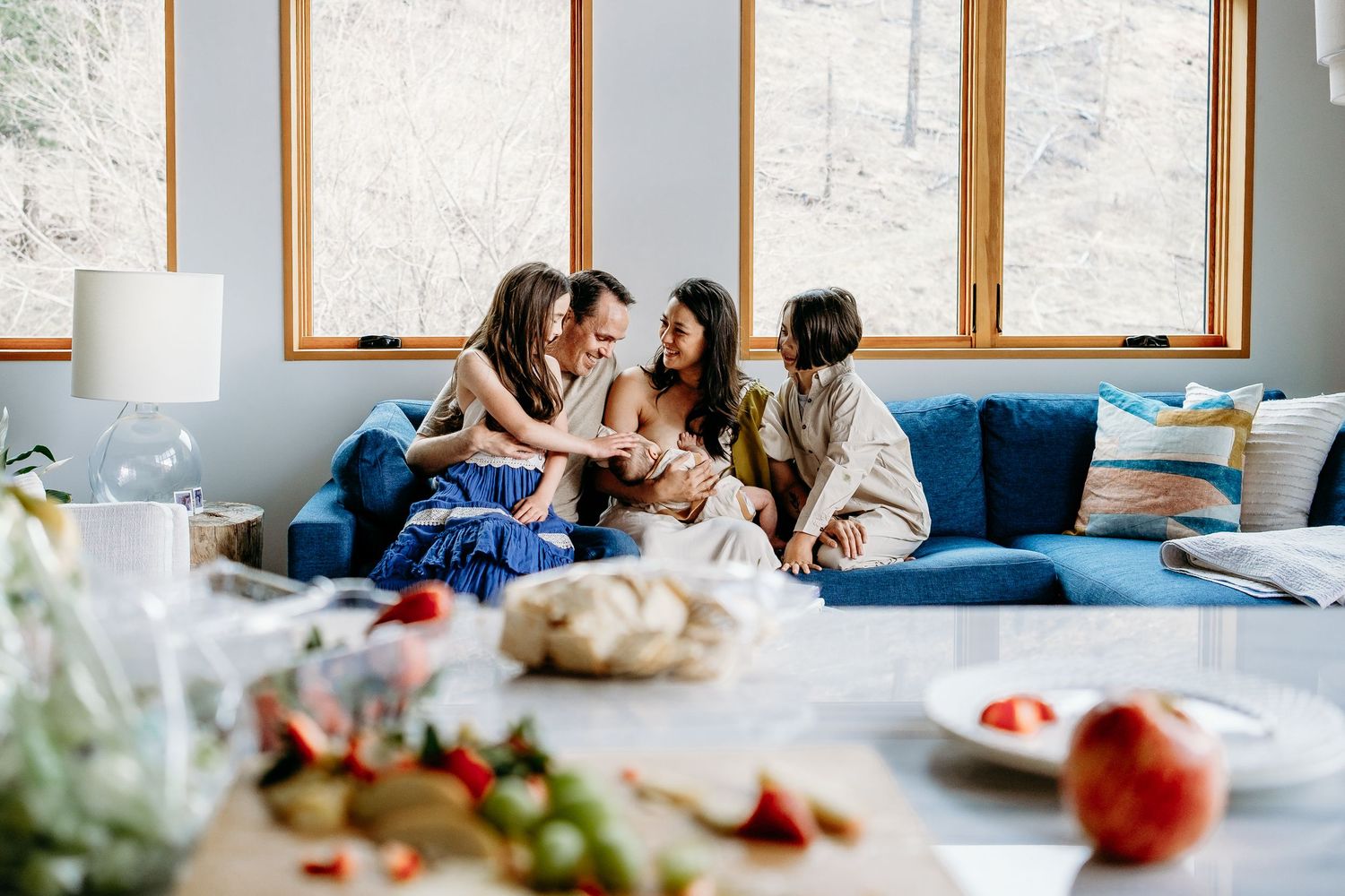 Denver family of daughter, father, mother breastfeeding newborn and son sitting on blue couch, counter filled with fruits in foreground.