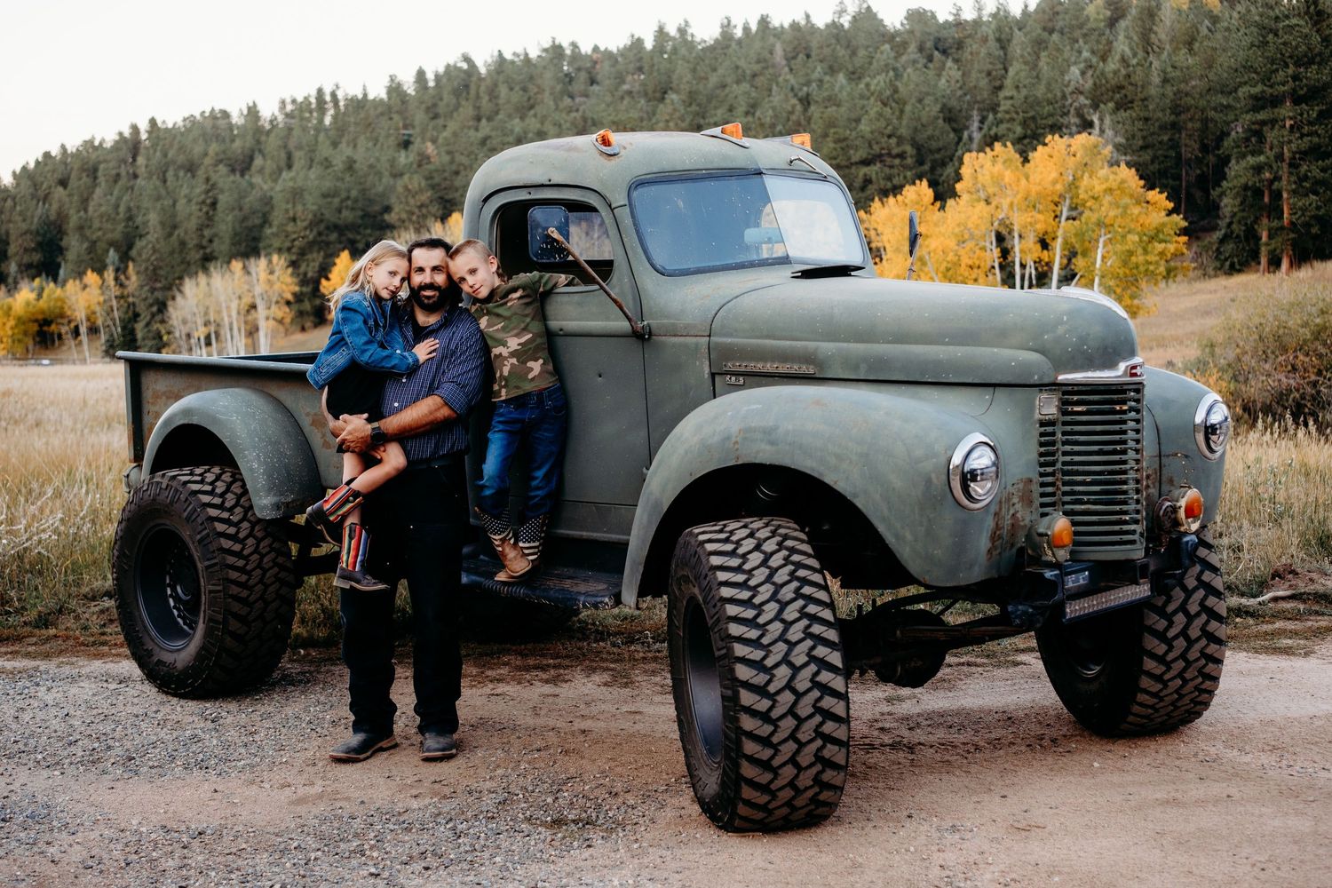Father holding daughter while son standings on floor boards of vintage green truck, all with serious expression.