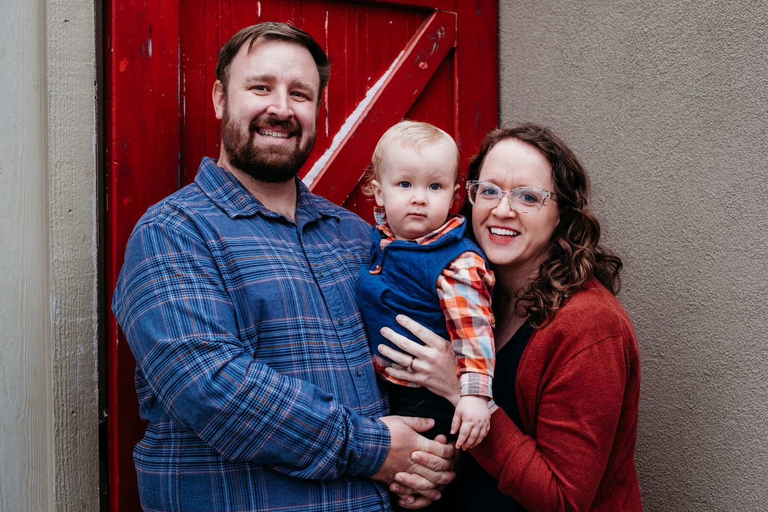 Father and mother holding toddler son in between them in front of red door in Denver, CO.