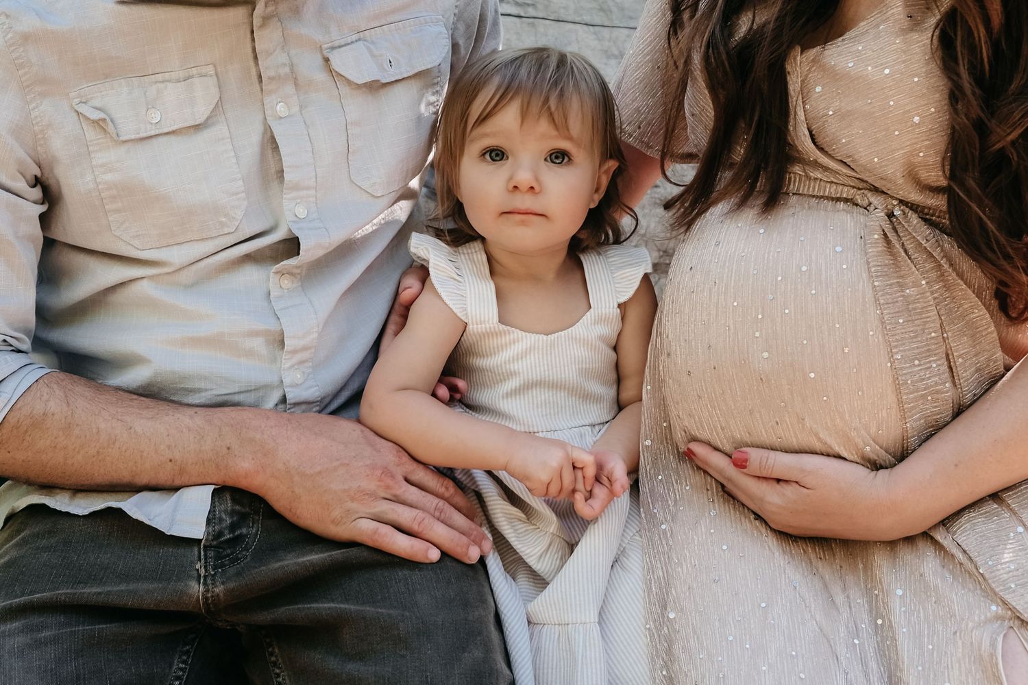 Toddler girl sitting between father and pregnant mother, looking thoughtfully toward camera.