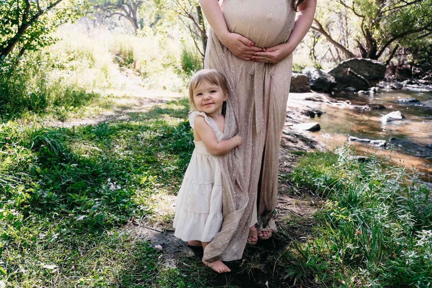 Close up of pregnant mother wearing champagne colored dress, toddler daughter hugging her leg, laughing.