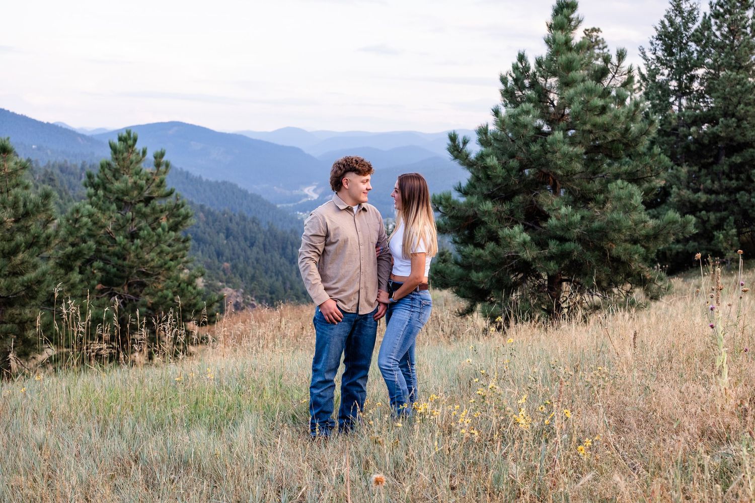 Young couple standing in field, mountains in background, both smiling at each other.