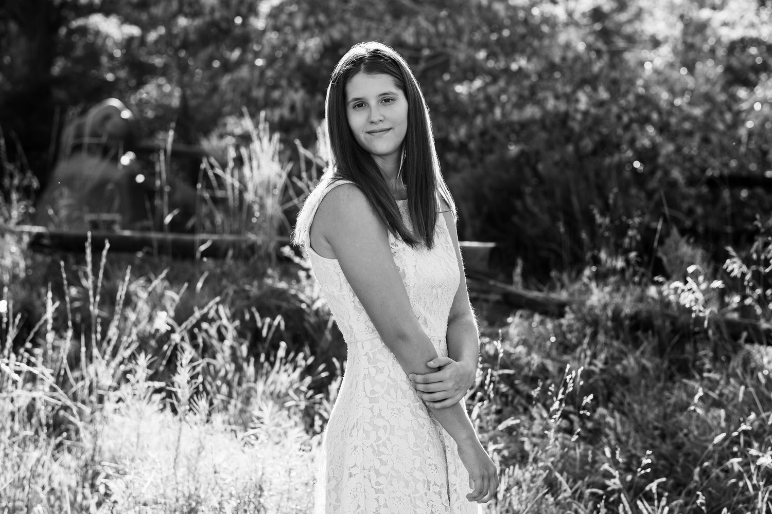 Black and white portrait of teen girl with thoughtful expression standing in tall grasses with trees in the background.