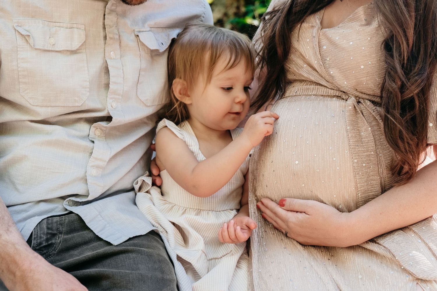 Toddler girl sitting between father and pregnant mother, gently touching mother's pregnant belly.