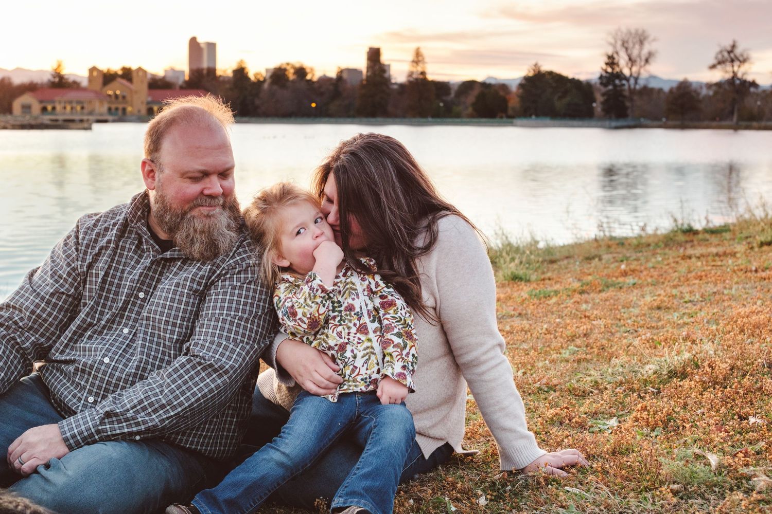 Father and mother sitting on grass, toddler sitting on mother's lap, Denver skyline, lake, City Park Pavilion in background.