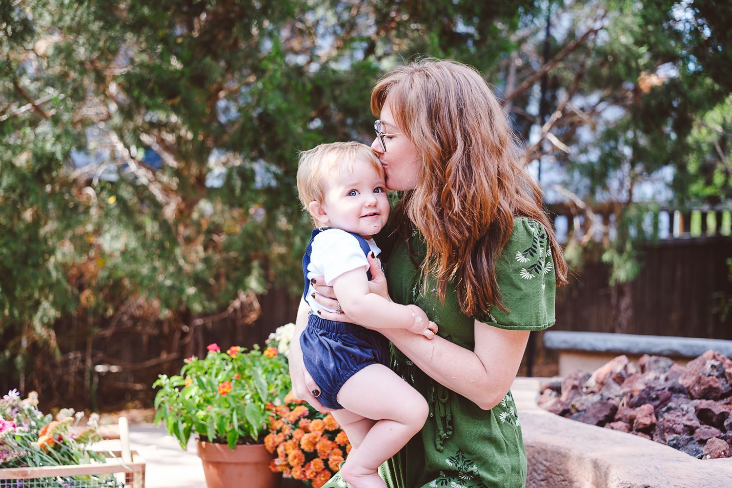 Mother kissing one year old son on his temple, in their backyard.