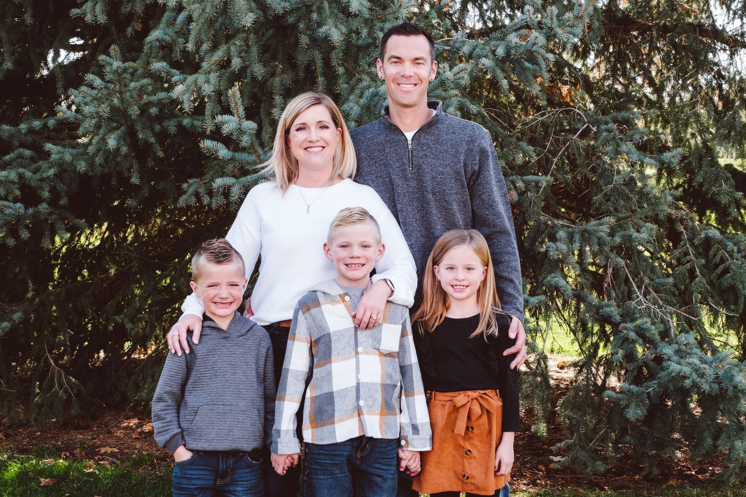 Family of five posing in front of evergreen trees in neighborhood park in Denver metro.