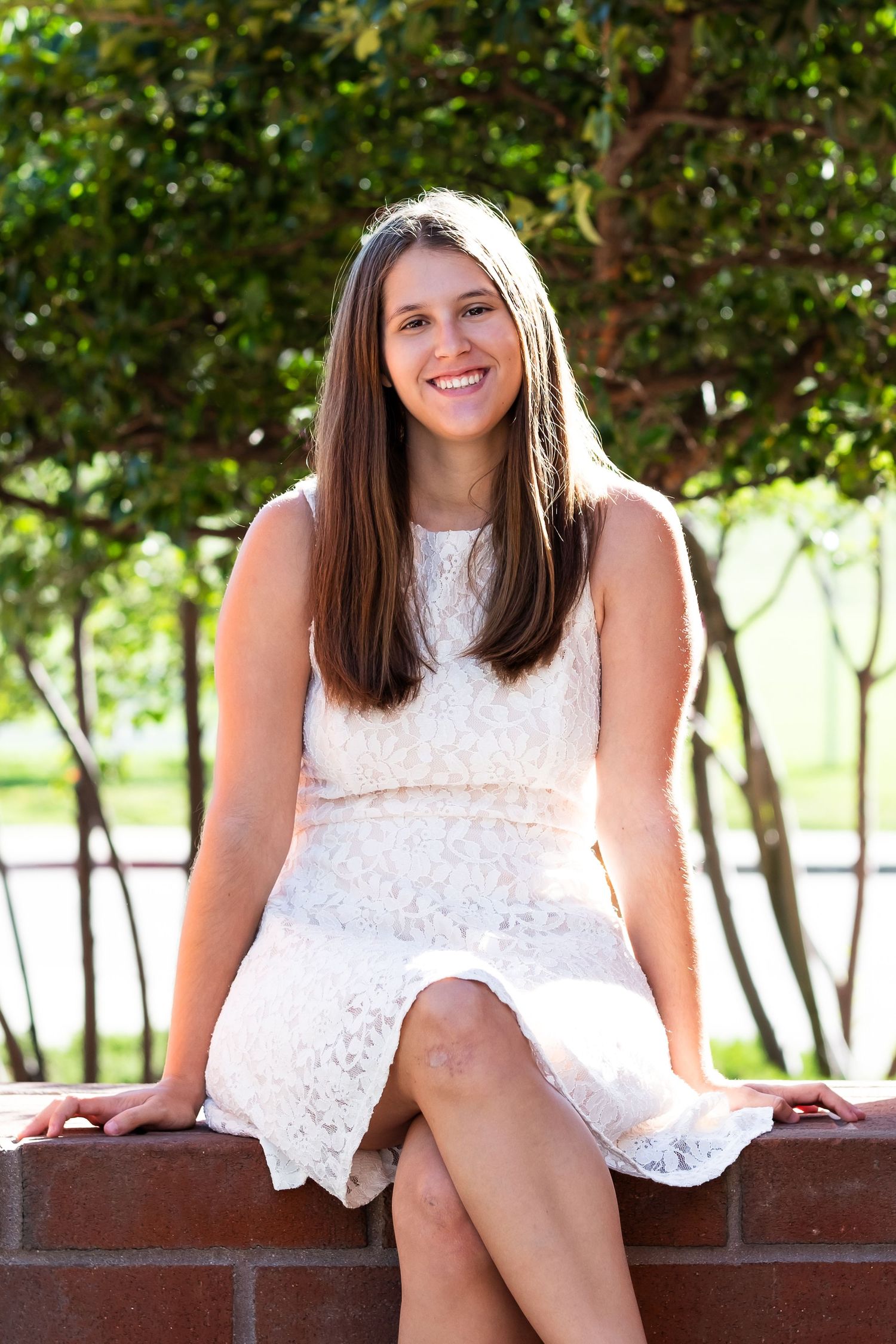 Senior portrait teen girl sitting on brick wall, wearing white dress.