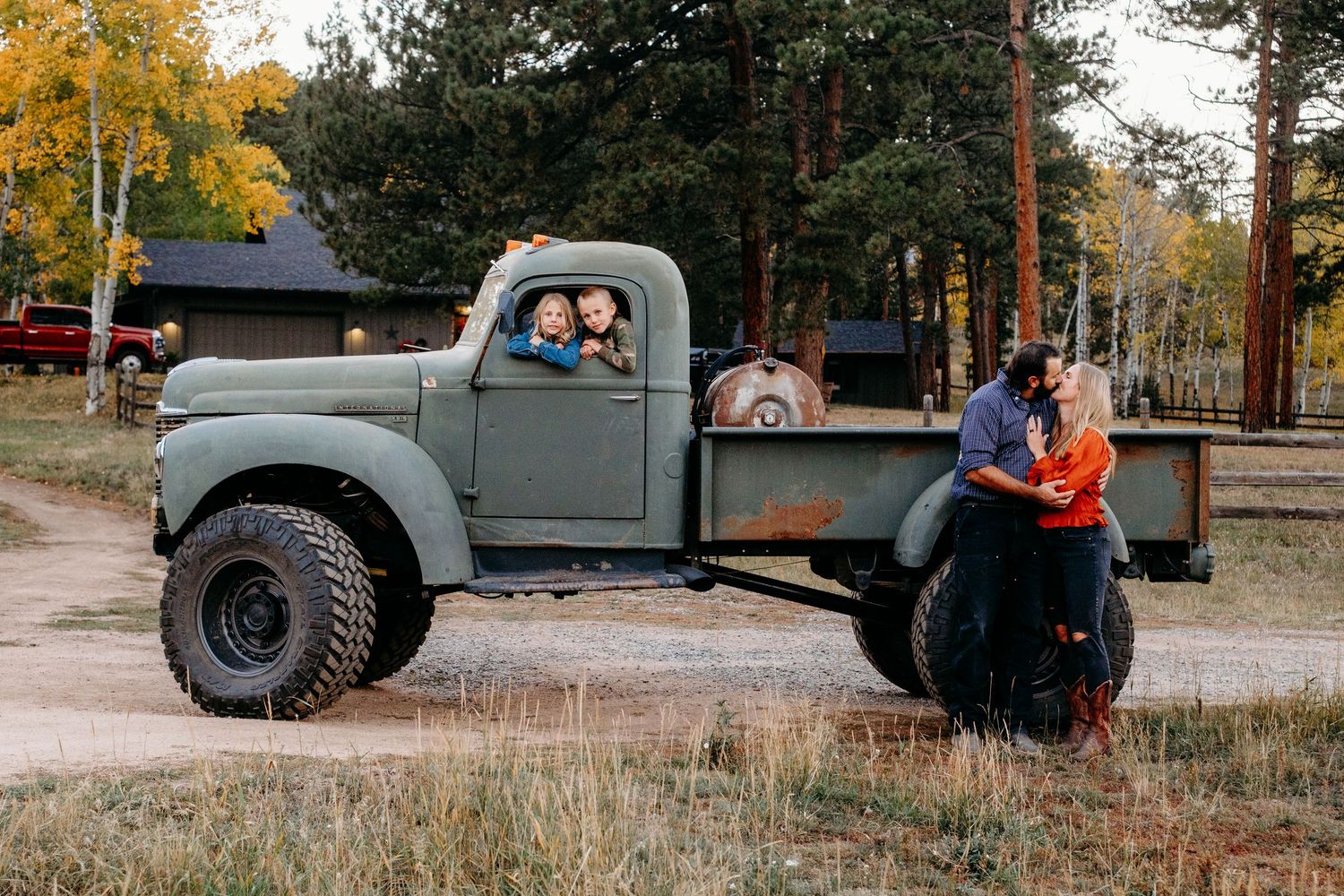 Brother and sister in front seat of large vintage green truck, mother and father standing by wheel well kissing.