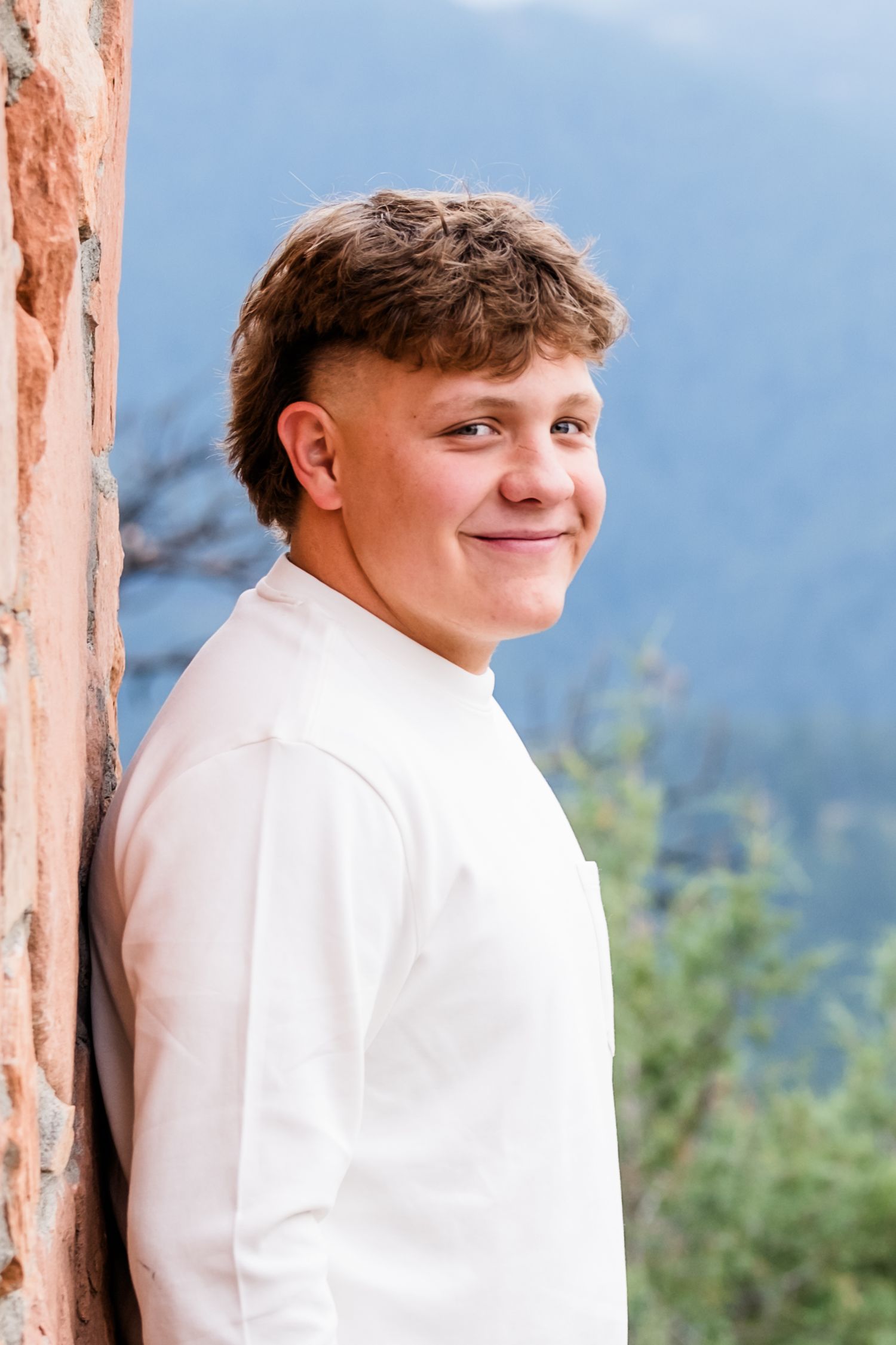 Traditional senior portrait, young man looking at camera and softly smiling while leaning against brick wall.