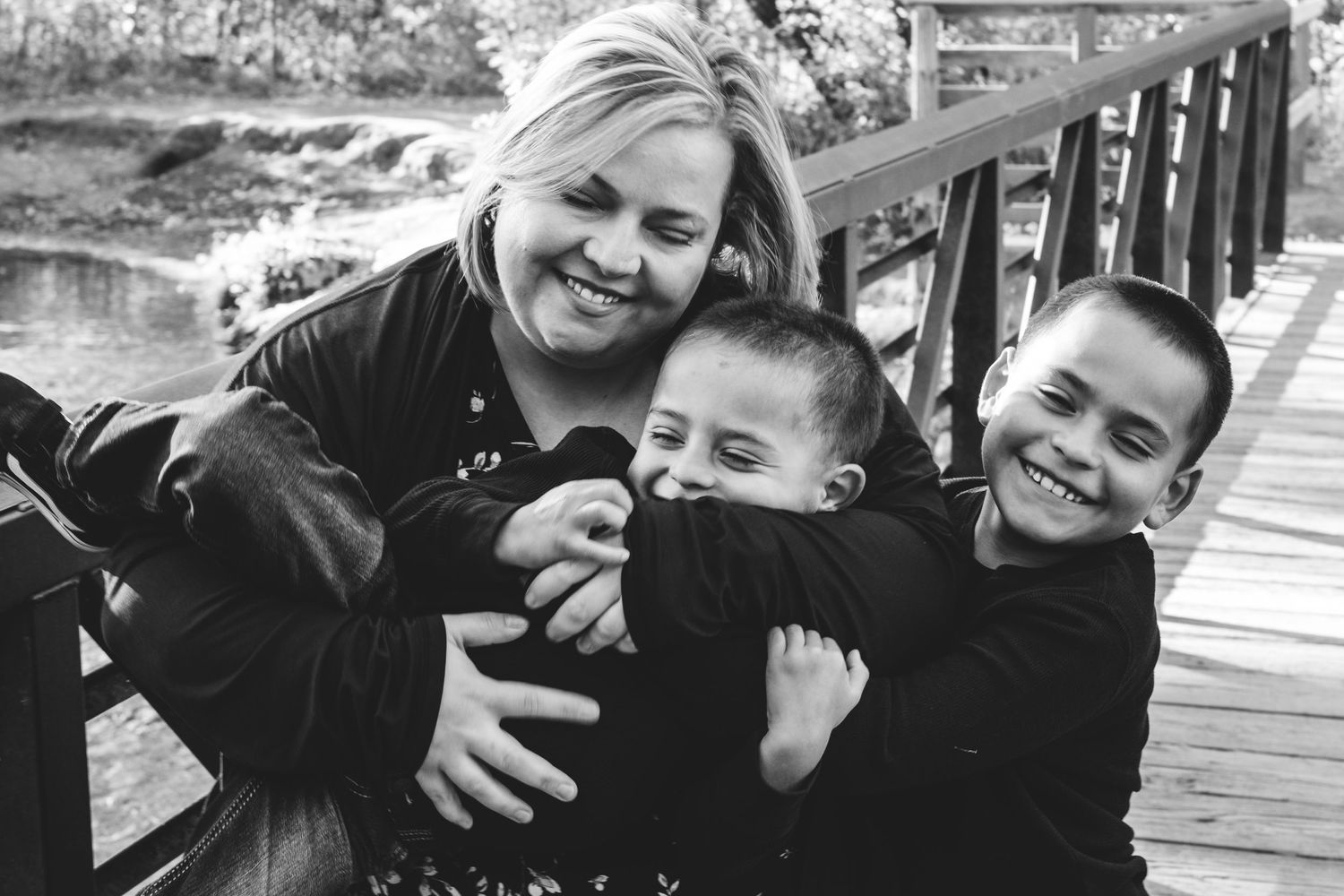 Candid photo of mother holding toddler son, older son hugging on mom, all three smiling widely, eyes closed.