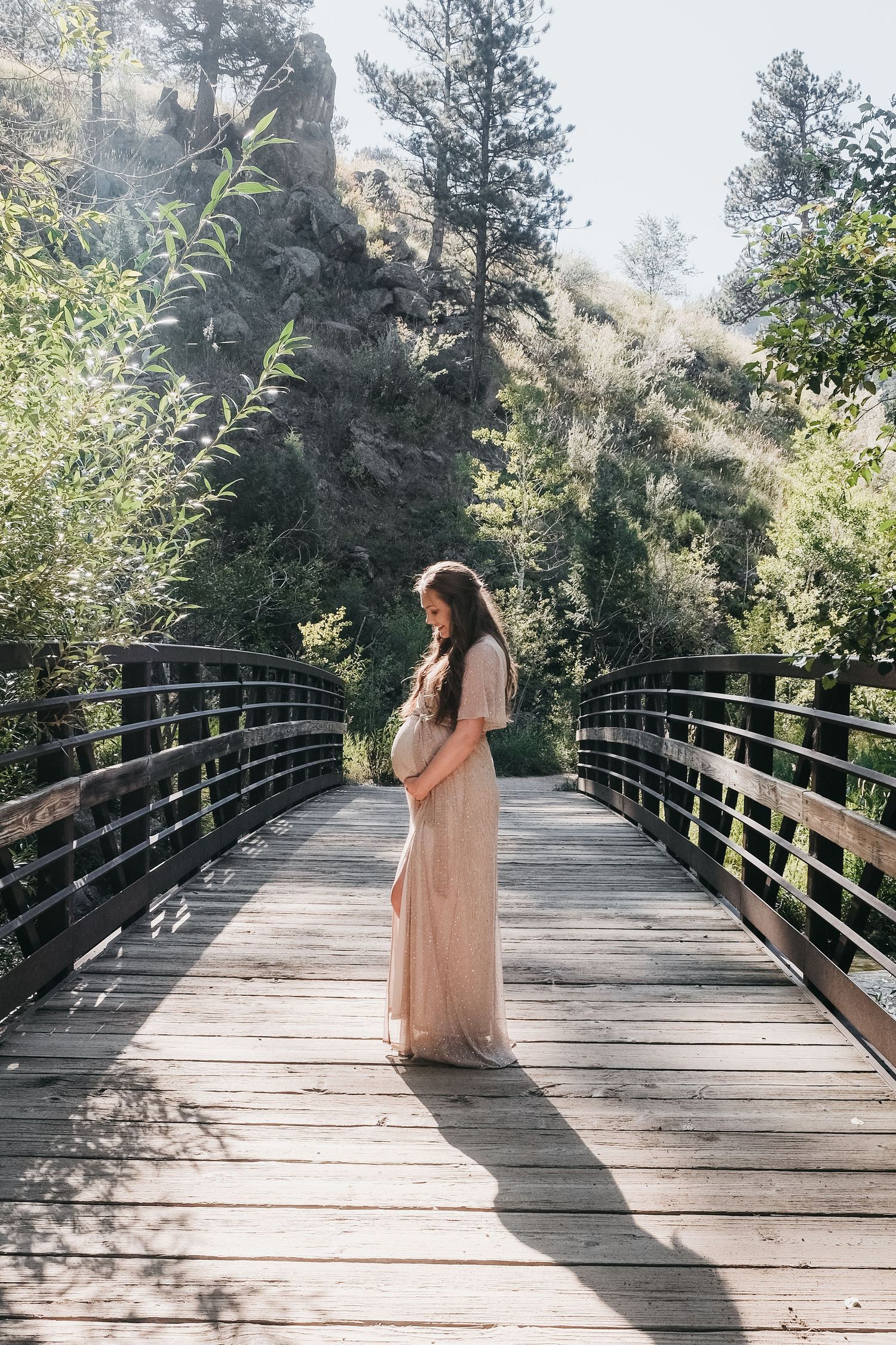 Profile of pregnant mother on wooden bridge with iron rails, sun bursting through tall green trees in the background.
