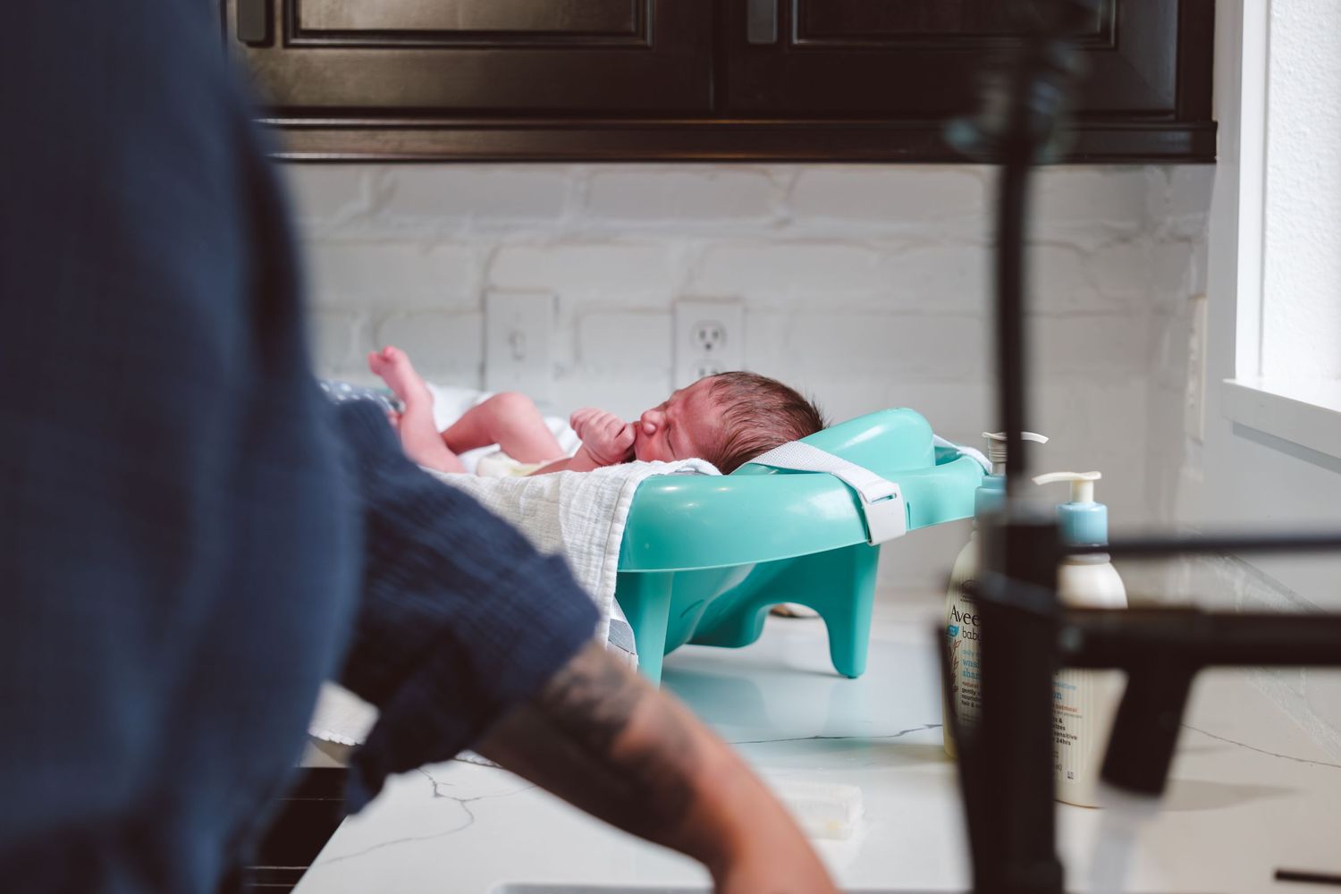 Newborn boy in infant bathtub on counter, mother washing hands in sink.