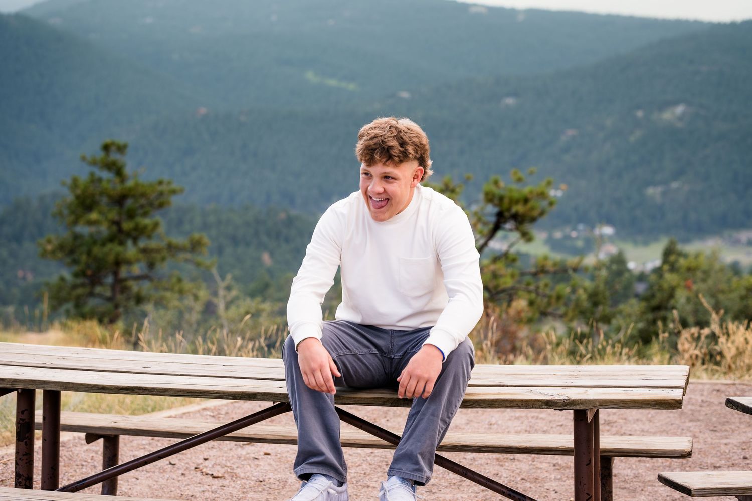 Senior portrait, senior laughing while sitting on a park picnic table, mountains in background.