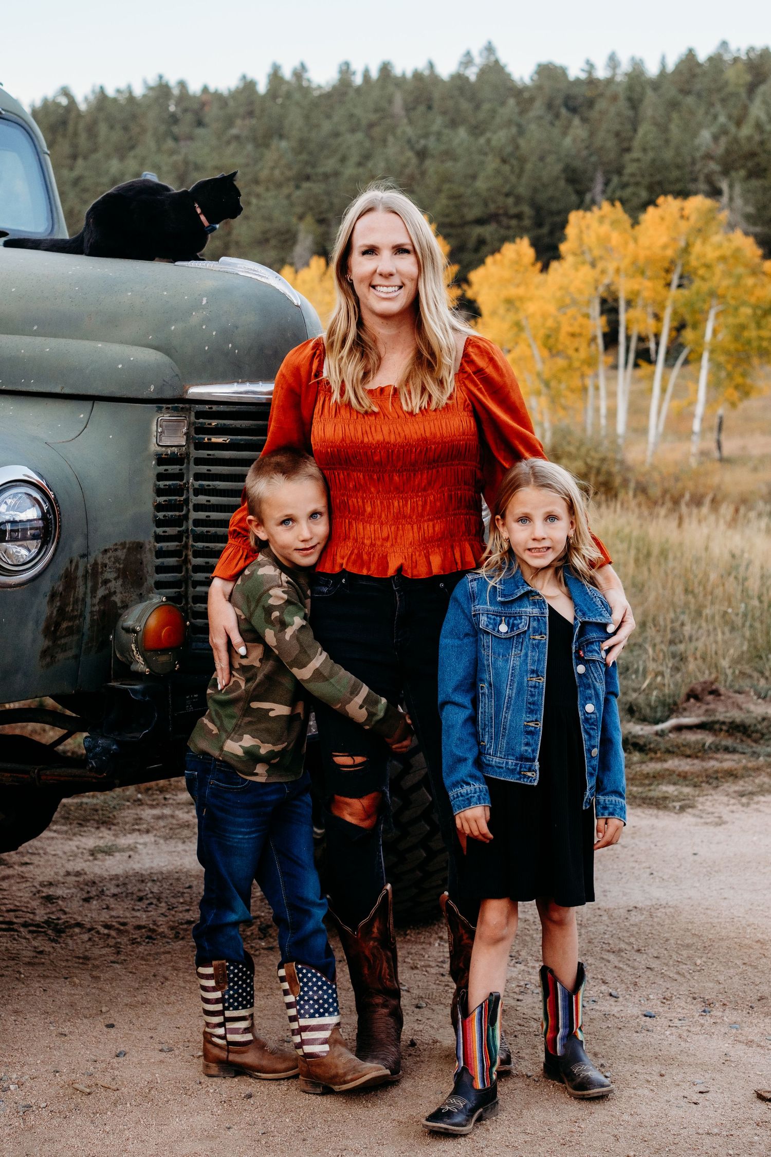 Mother, son and daughter stand in front of vintage green truck, black cat perched on hood of truck.