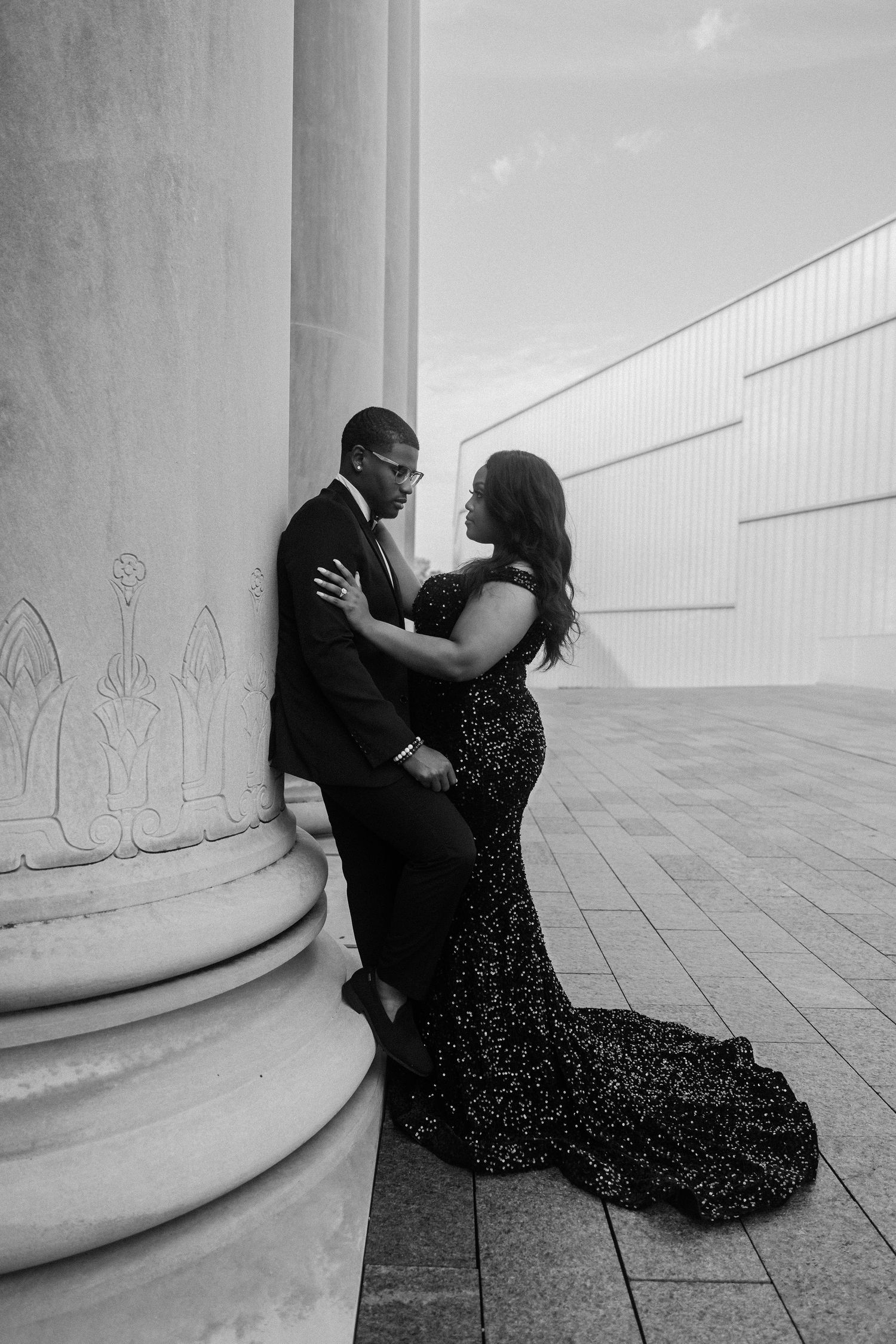 Couple in formal wear poses against classic column in black and white.