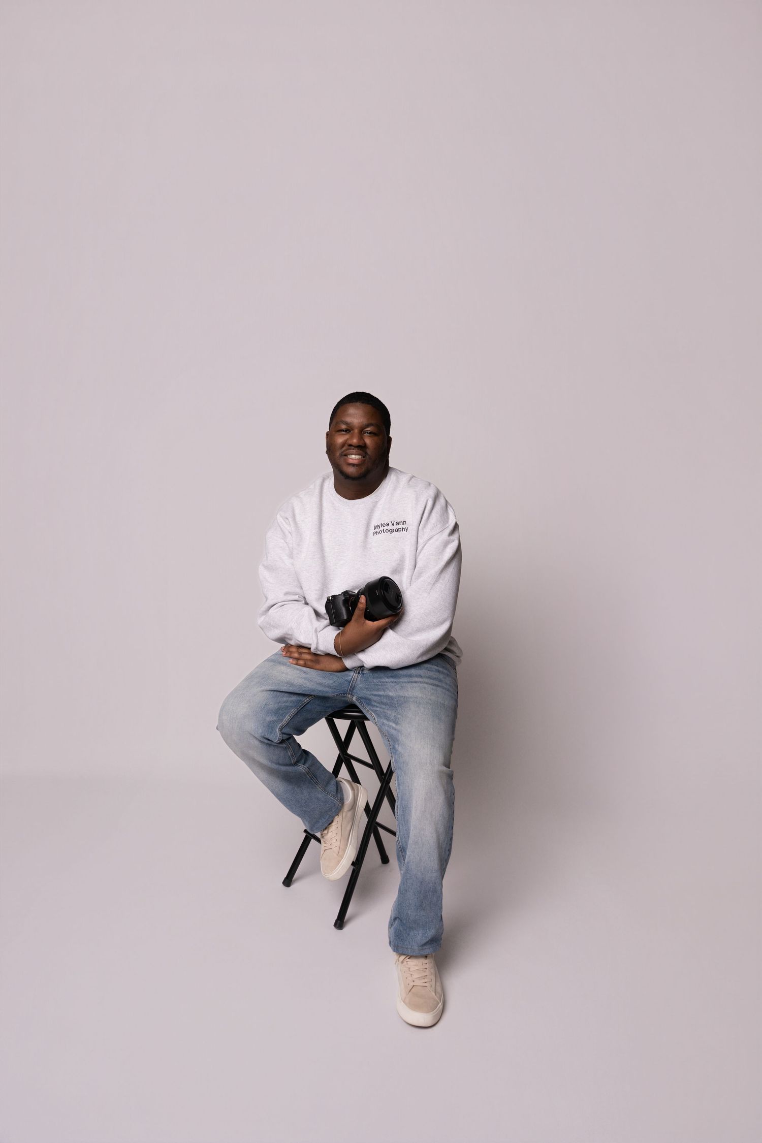 A photographer wearing a white sweatshirt and jeans sits casually on a stool against a light gray backdrop while holding a camera.