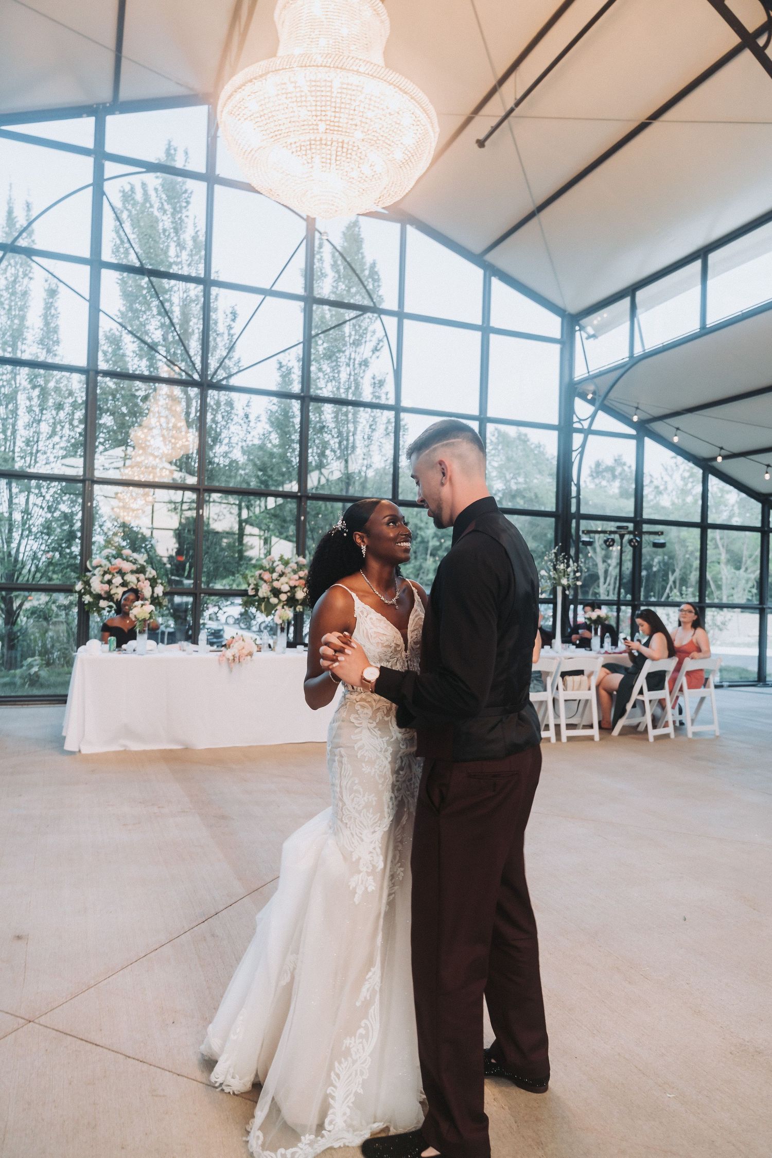 Wedding couple dancing in elegant glass-walled venue with chandelier overhead.