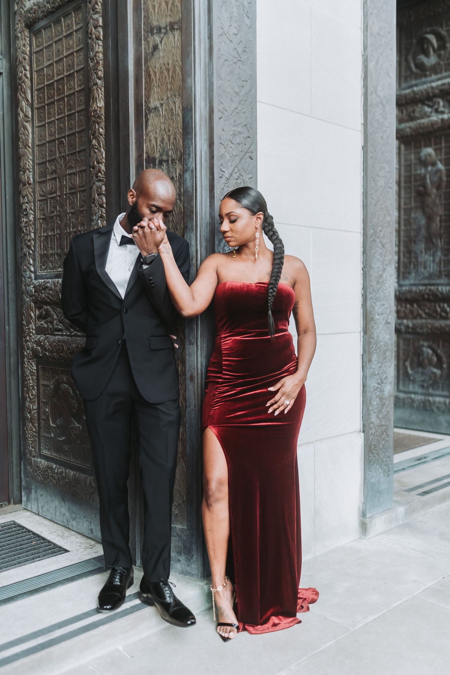 Elegant couple poses against stone building exterior, with woman in red velvet gown and man in classic tuxedo.