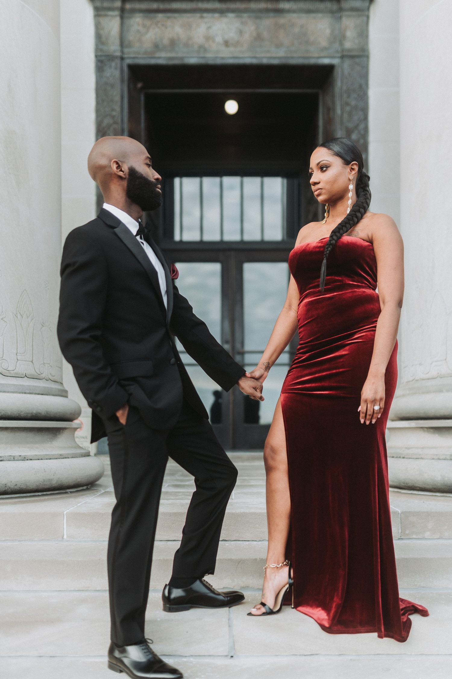 Fashionable couple poses by classical columns, with woman in red velvet gown and man in black suit.