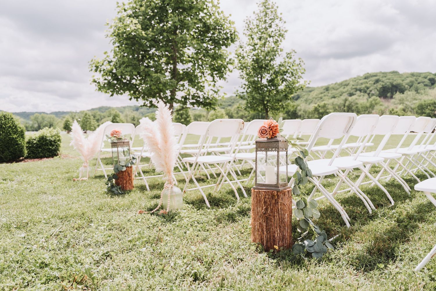White chairs set up for an outdoor wedding ceremony with rustic wooden lantern stands and floral decorations.
