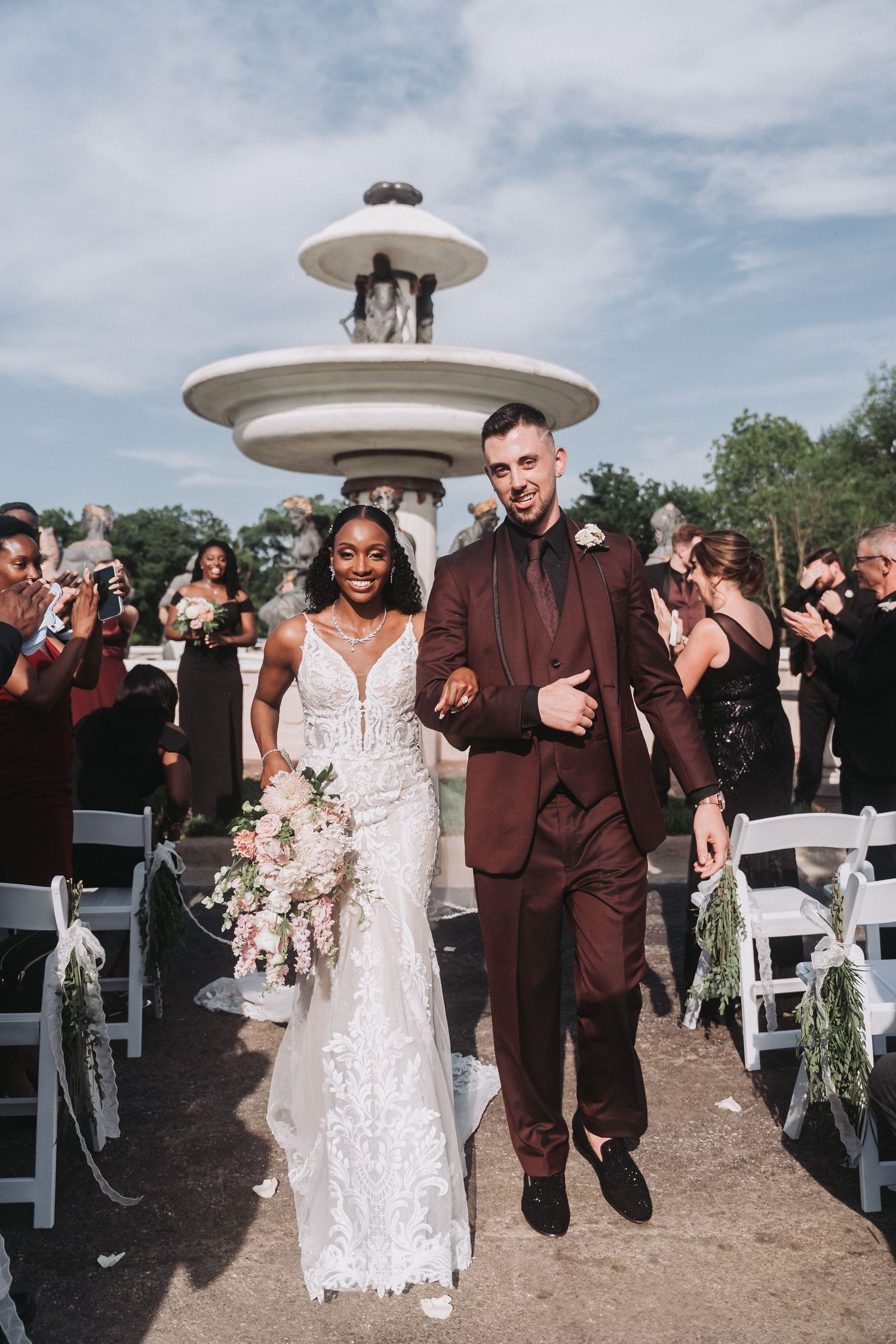 Wedding couple walking down aisle past elegant fountain while guests cheer.