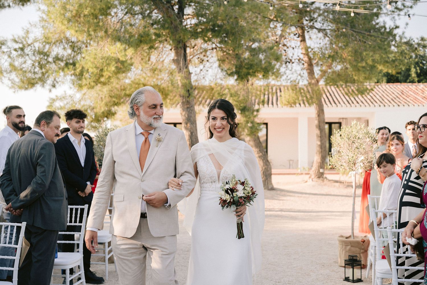 A sunny outdoor wedding ceremony procession along a garden path lined with trees at a Mediterranean villa location.