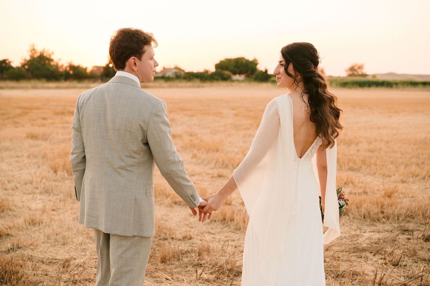Newlywed couple holding hands in golden field at sunset wearing gray suit and white wedding dress.