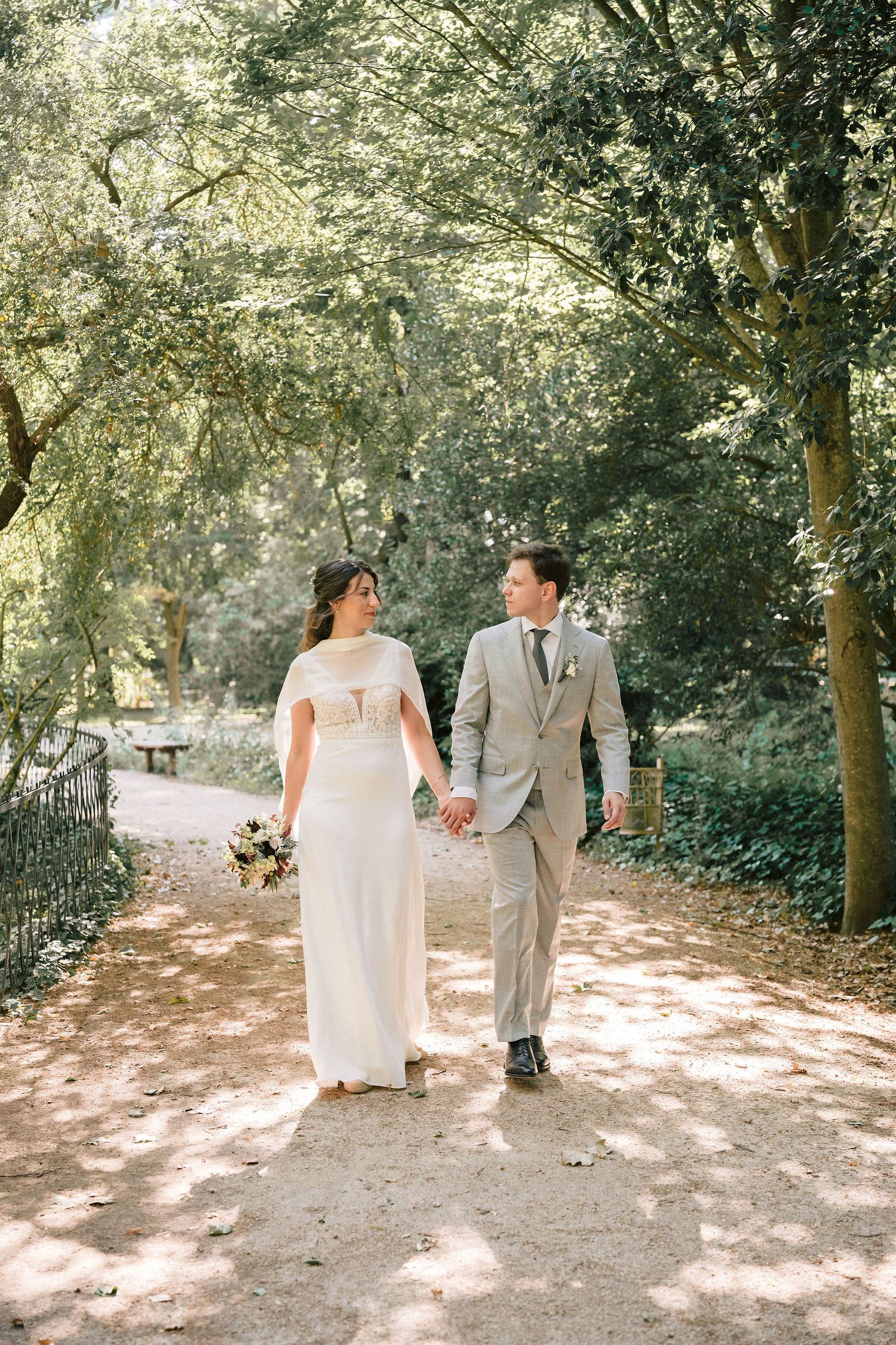 Romantic wedding couple walks along dappled sunlit garden path on their special day.