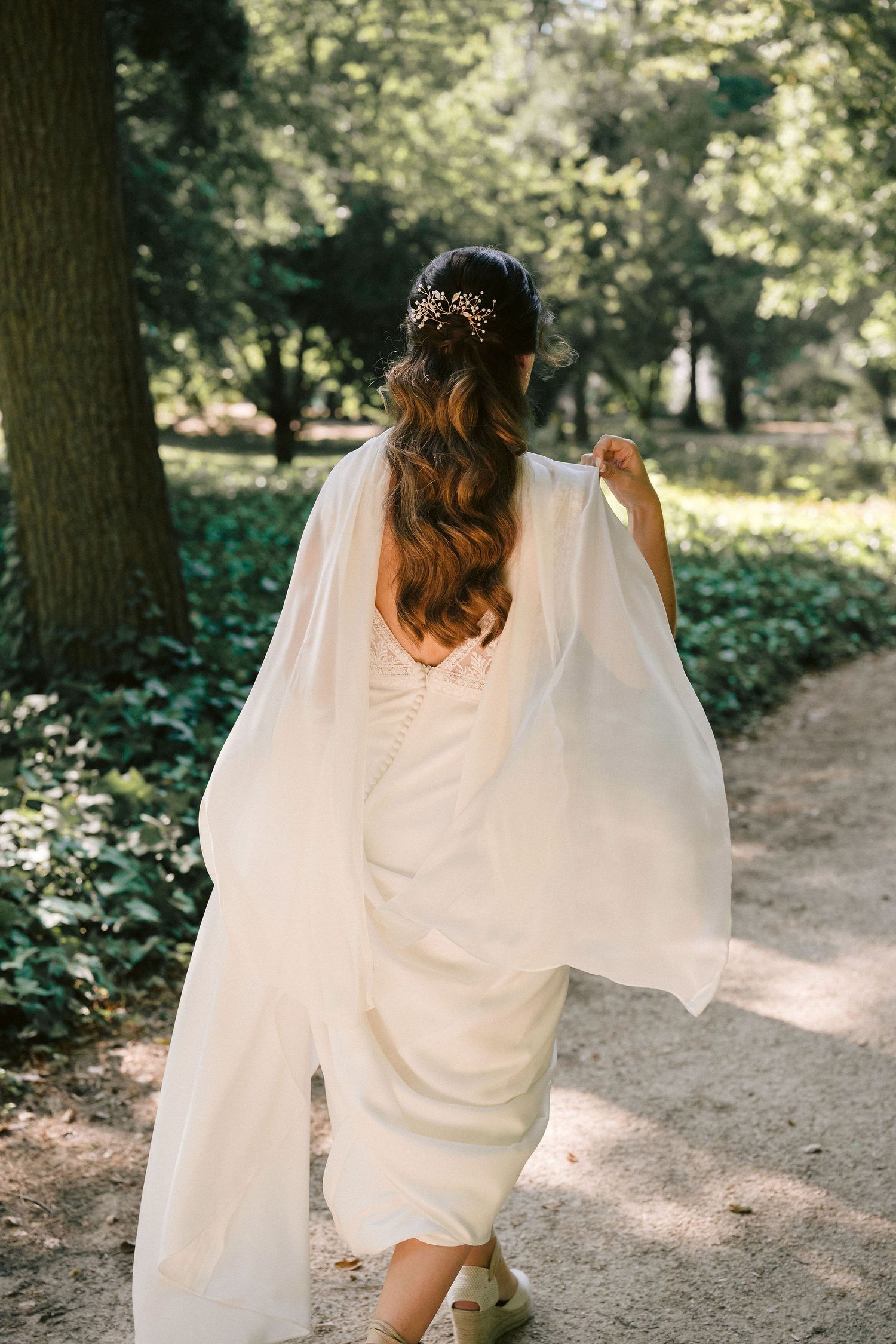 A bride with flowing wavy hair and sparkly headpiece wears a white dress with flowing sleeves walking on a forest path.