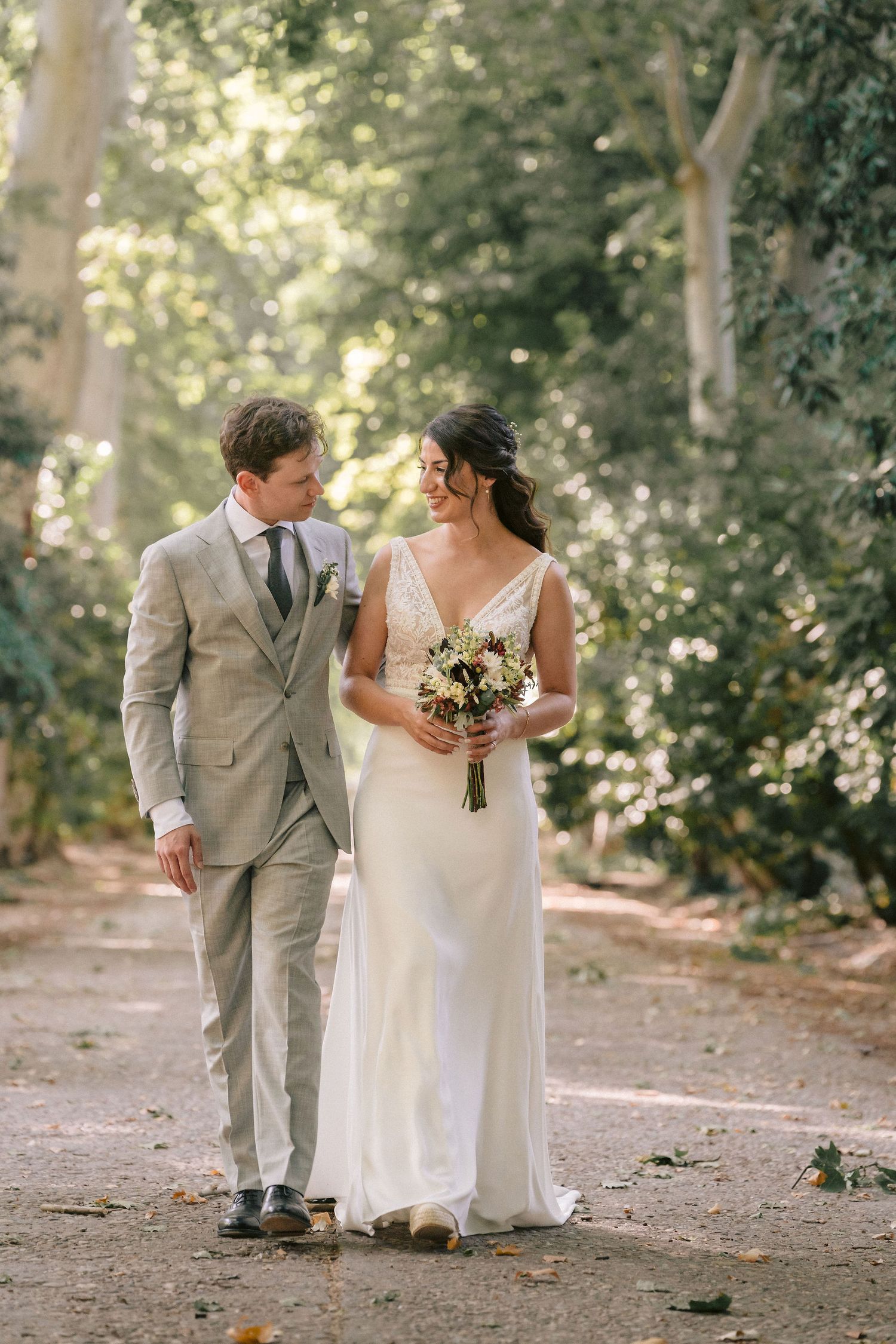 A wedding couple walking together on a tree-lined path in grey suit and white dress while holding a floral bouquet.