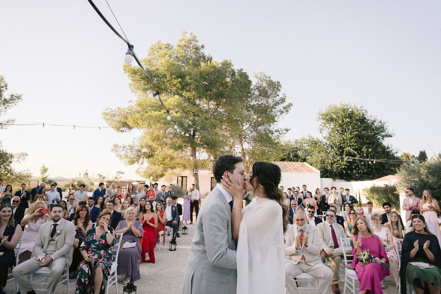 A couple shares a romantic kiss during their outdoor wedding ceremony as guests look on at a sunlit venue.