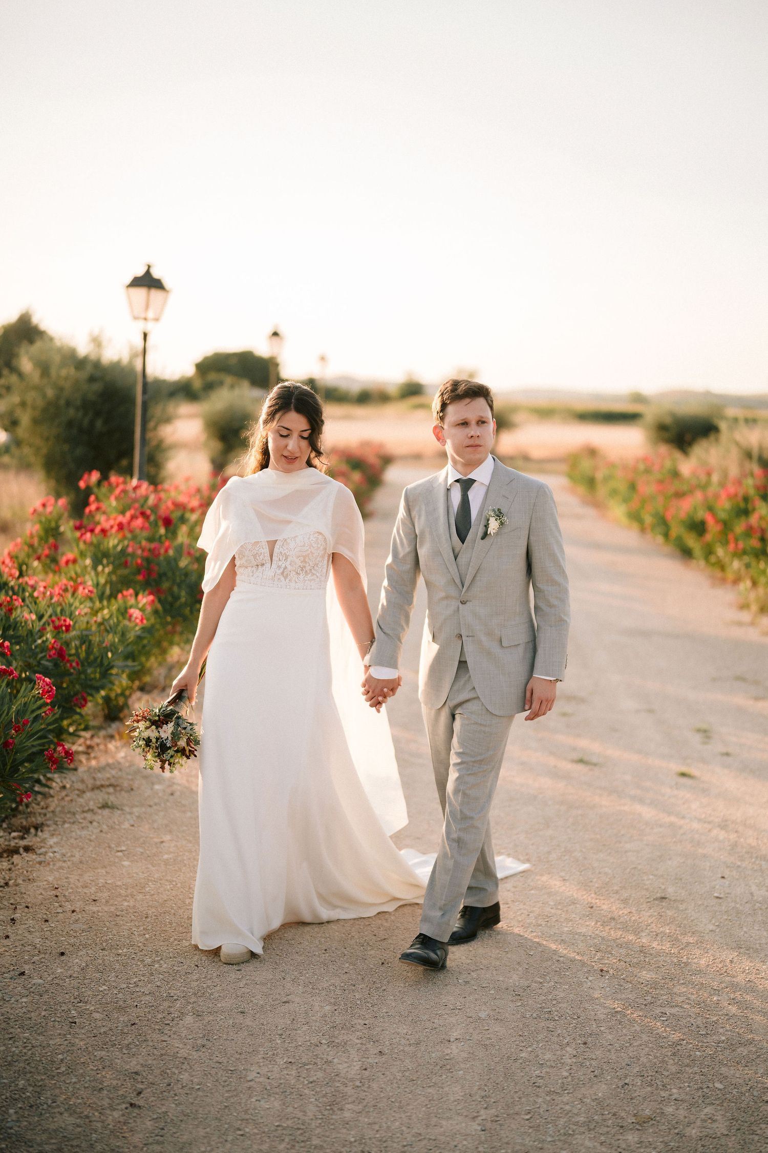 A bride and groom walk hand in hand on a path lined with red flowers at sunset during a romantic wedding photo session.