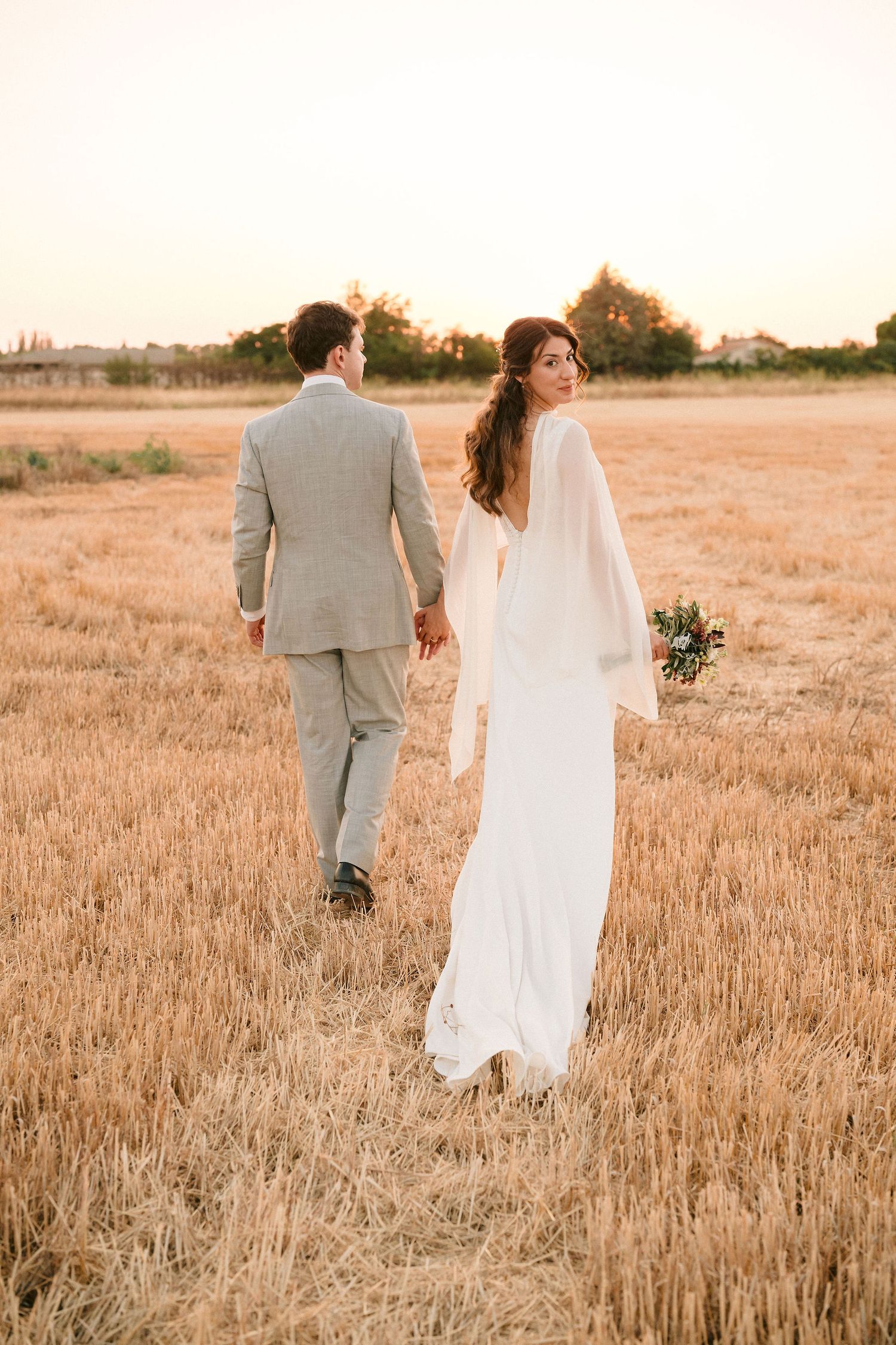 A newlywed couple walks through a golden wheat field at sunset holding hands in romantic wedding attire.