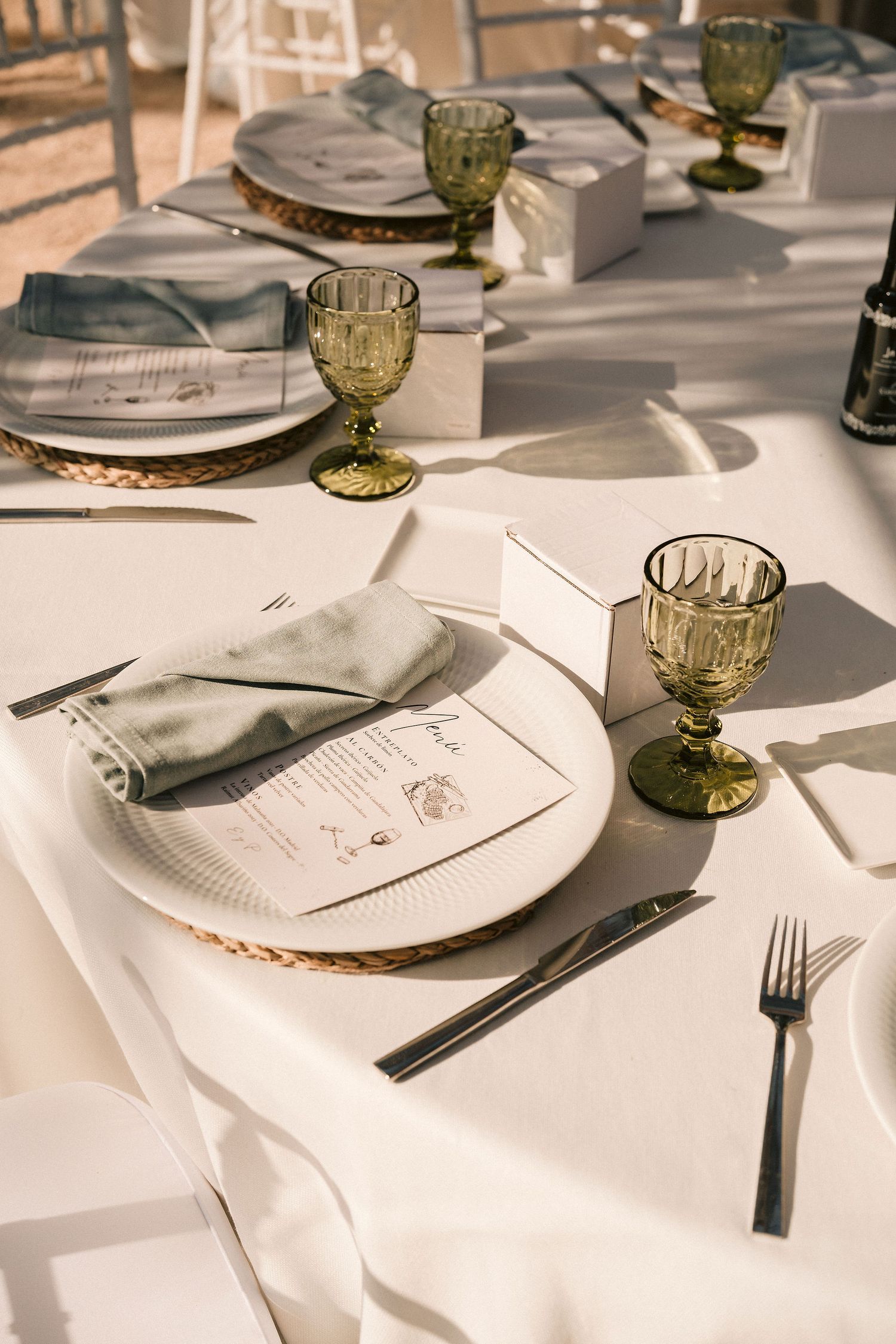 White tablecloth place settings with gold-rimmed plates, green vintage glassware, and printed menus at an elegant dinner.