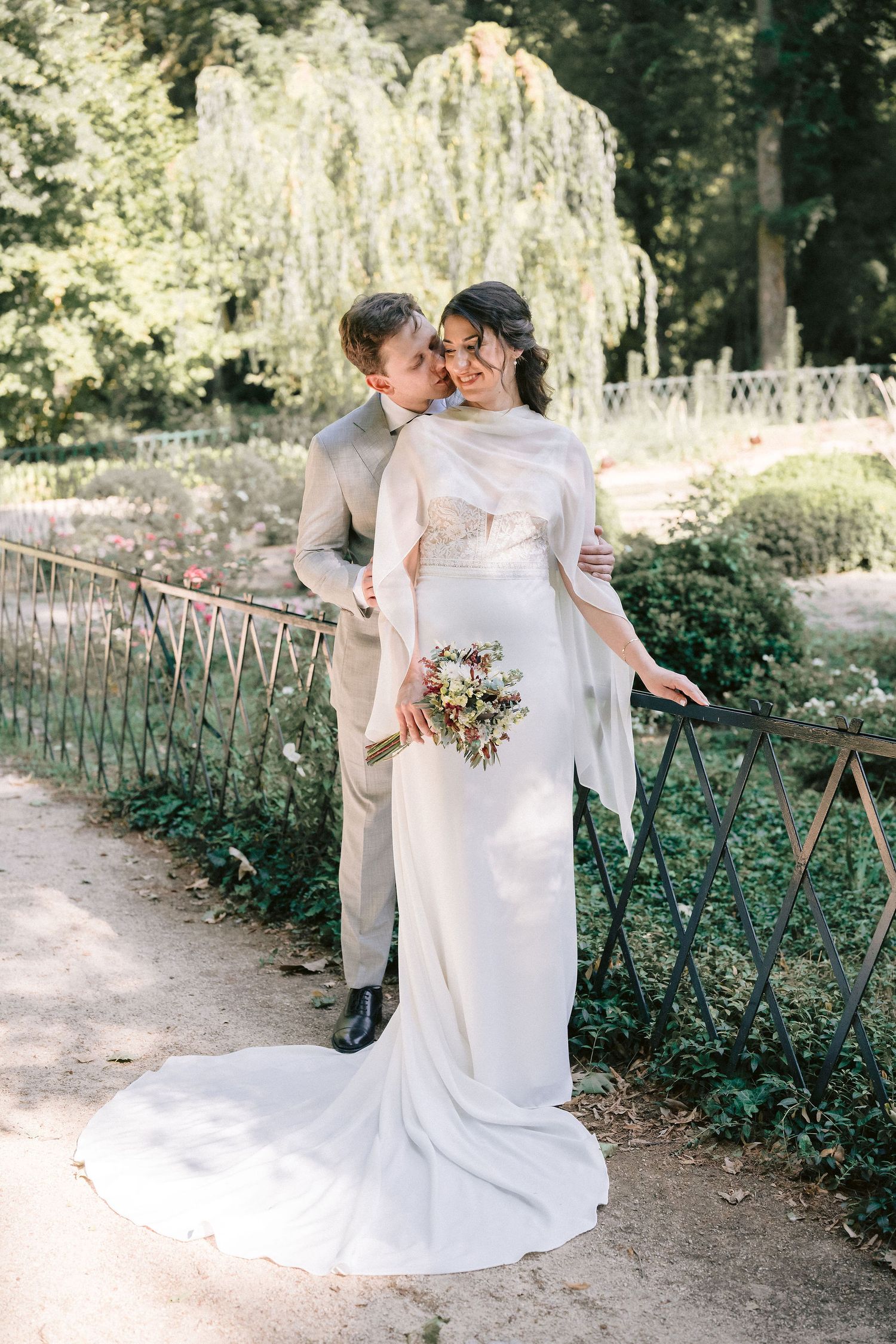 A couple in wedding attire embraces on a garden pathway surrounded by lush greenery and flowers on their special day.