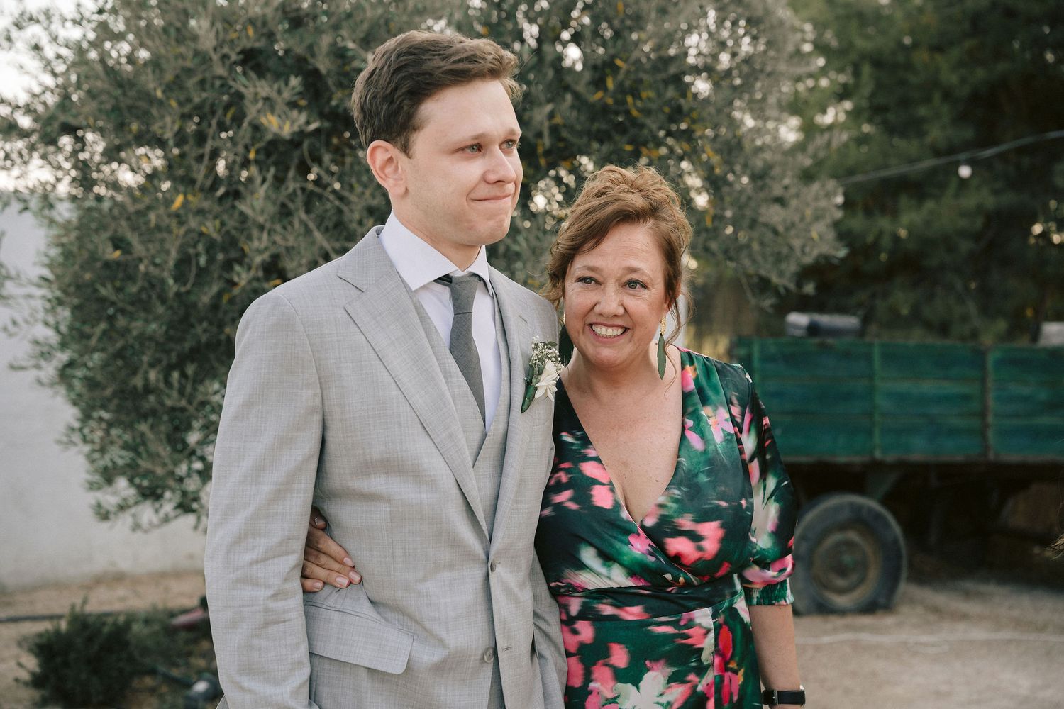A couple in wedding attire poses outdoors, with the bride wearing a floral dress and groom in a light gray suit.